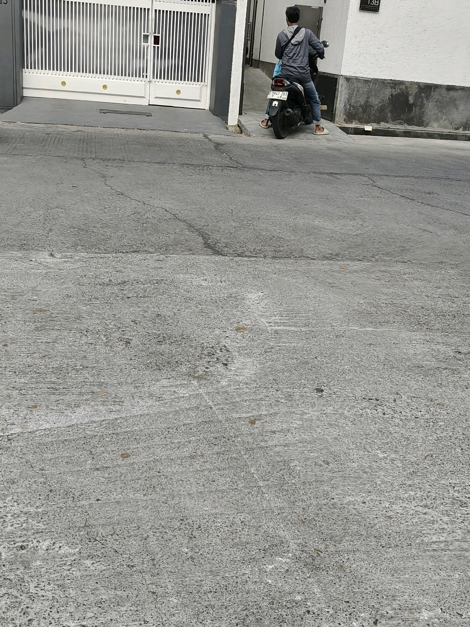 The image shows a person on a motorcycle parked on a concrete surface. The motorcycle is dark in color with a license plate that reads "D 5842 ZB1". The rider, wearing a grey hoodie and blue jeans, is facing away from the camera and appears to be looking at a white gate with vertical slats. The gate is part of a building with a white wall. Behind the gate, there is a darker grey wall with vertical lines. The scene appears to be an outdoor urban environment, likely a street or driveway in Bandung City, Indonesia, given the license plate format. The concrete surface is worn and cracked, indicating it is an older surface.