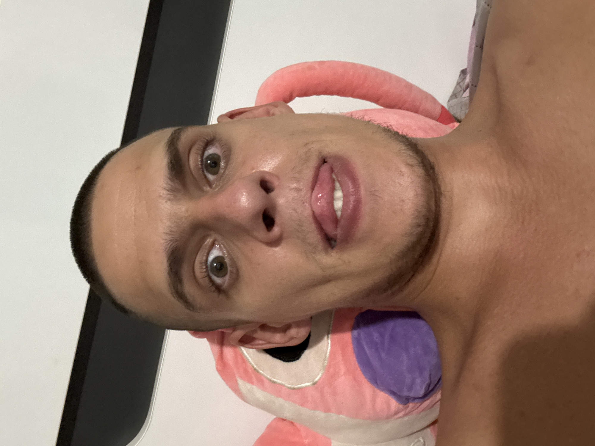 A close-up, selfie-style shot features a shirtless person with a buzz cut, looking directly at the camera with wide eyes and their tongue sticking out. The person is leaning against a large, pink stuffed animal, possibly a character from a cartoon, which has a purple section near the person's head. The background appears to be a wall with a dark horizontal band across the top. The lighting suggests an indoor setting, possibly a bedroom. No specific location in Sofia, Bulgaria can be determined from the visual information provided.