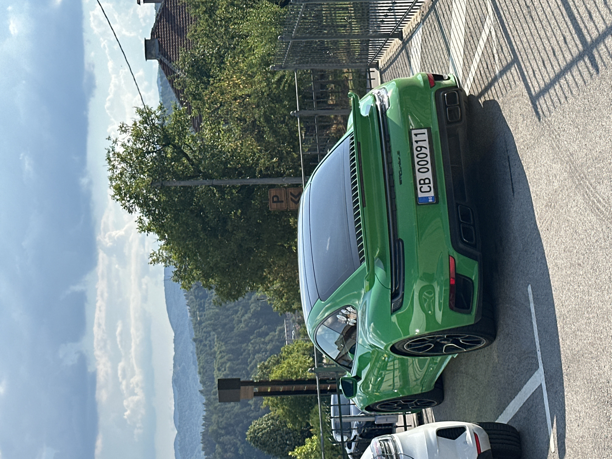 A vibrant green Porsche 911 Turbo S is parked on an asphalt surface in Kerenite, Bulgaria. The car is positioned at an angle, with its rear facing the viewer. The license plate reads "CB 000911". Adjacent to the Porsche, a white SUV is partially visible on the left. In the background, a black metal fence separates the parking area from a wooded hillside. Trees with lush green foliage dominate the midground, and distant hills are visible under a sky with scattered clouds. The scene is illuminated by sunlight, casting shadows from the fence onto the parking lot. A traffic sign with the letter "P" is visible on a pole to the left of the Porsche.
