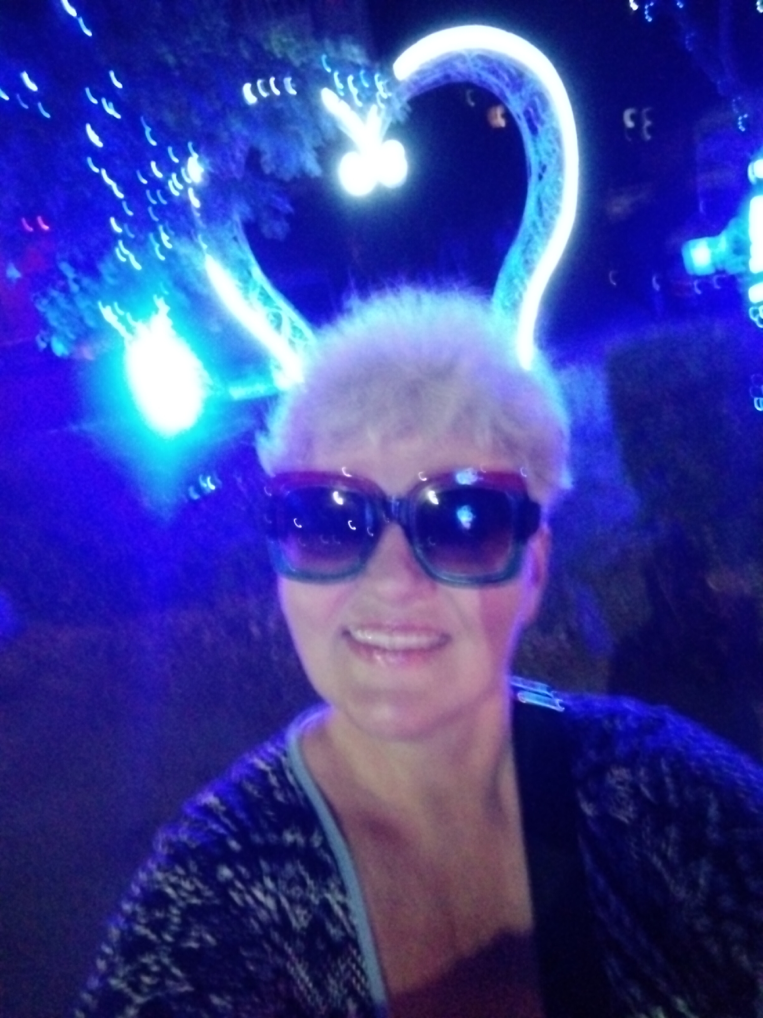 A woman with short, light-colored hair and large, dark-rimmed sunglasses smiles at the camera. She is wearing a dark patterned sweater with a light blue trim. She is illuminated by bright blue lights, and behind her, a large, luminous heart shape made of lights is visible against a dark background. The overall scene suggests an outdoor nighttime setting, possibly an event or celebration. The image is slightly blurry and has a bokeh effect from the lights. The photo was taken in Horishni Plavni, Ukraine.