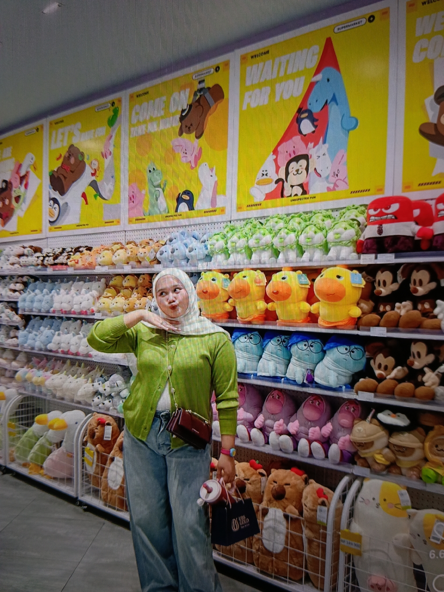 The image shows a young woman wearing a hijab and a green cardigan standing in a toy store. She is posing for the camera with her hand near her face. The store is filled with shelves of plush toys, including stuffed animals, cartoon characters, and other novelty items. In the background, there are large posters with colorful illustrations and text. The woman is holding a bag and a water bottle. The store appears to be well-lit and organized. The location is specified as the city of Raha, Indonesia.