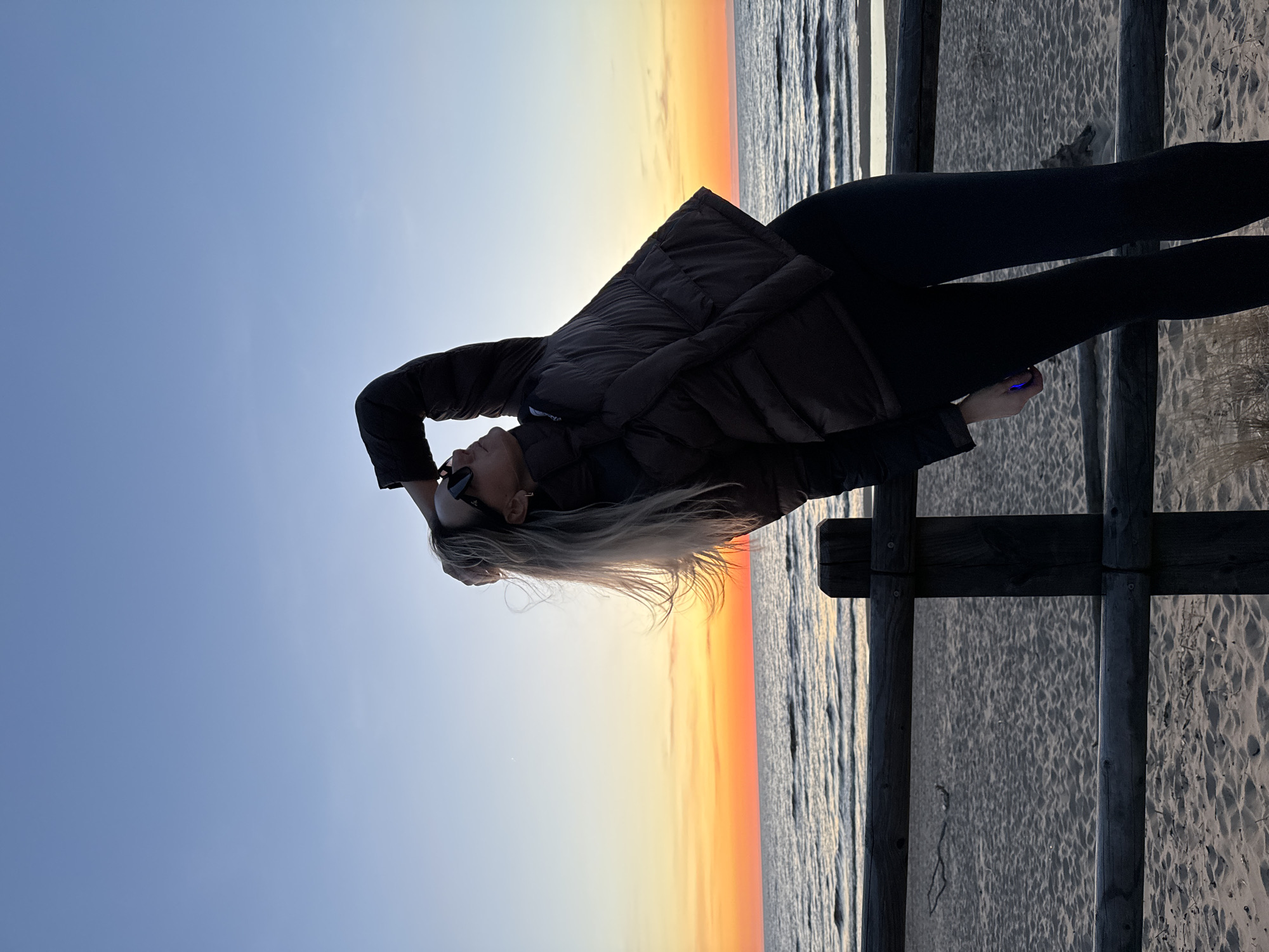 The image shows a woman with blonde hair wearing sunglasses, a dark puffer jacket, and black leggings, standing on a beach in Garciems, Latvia. She is positioned next to a wooden fence. The background reveals a sunset over the ocean, transitioning from orange to blue. The beach sand is visible through the fence. The woman has one hand on her head and is holding a small object in her other hand.