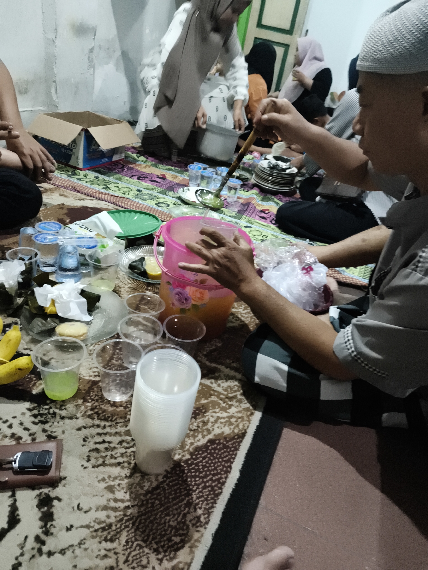 The image depicts a casual gathering of people, possibly a family, sitting on the floor in what appears to be a room in Barengkrajan, Indonesia. The setting is informal, with people seated on a patterned rug. A variety of food and drink items are spread across the rug, including a large pink bucket filled with a yellowish liquid, plastic cups, plates with food, and bananas. Several individuals are visible, some wearing traditional Indonesian attire. One person in the foreground is holding a plastic bag filled with something red. Another person is using a wooden utensil, perhaps to stir the drink. The environment appears to be indoors, with plain walls and a glimpse of a doorway in the background. The overall impression is one of a communal meal or social event.