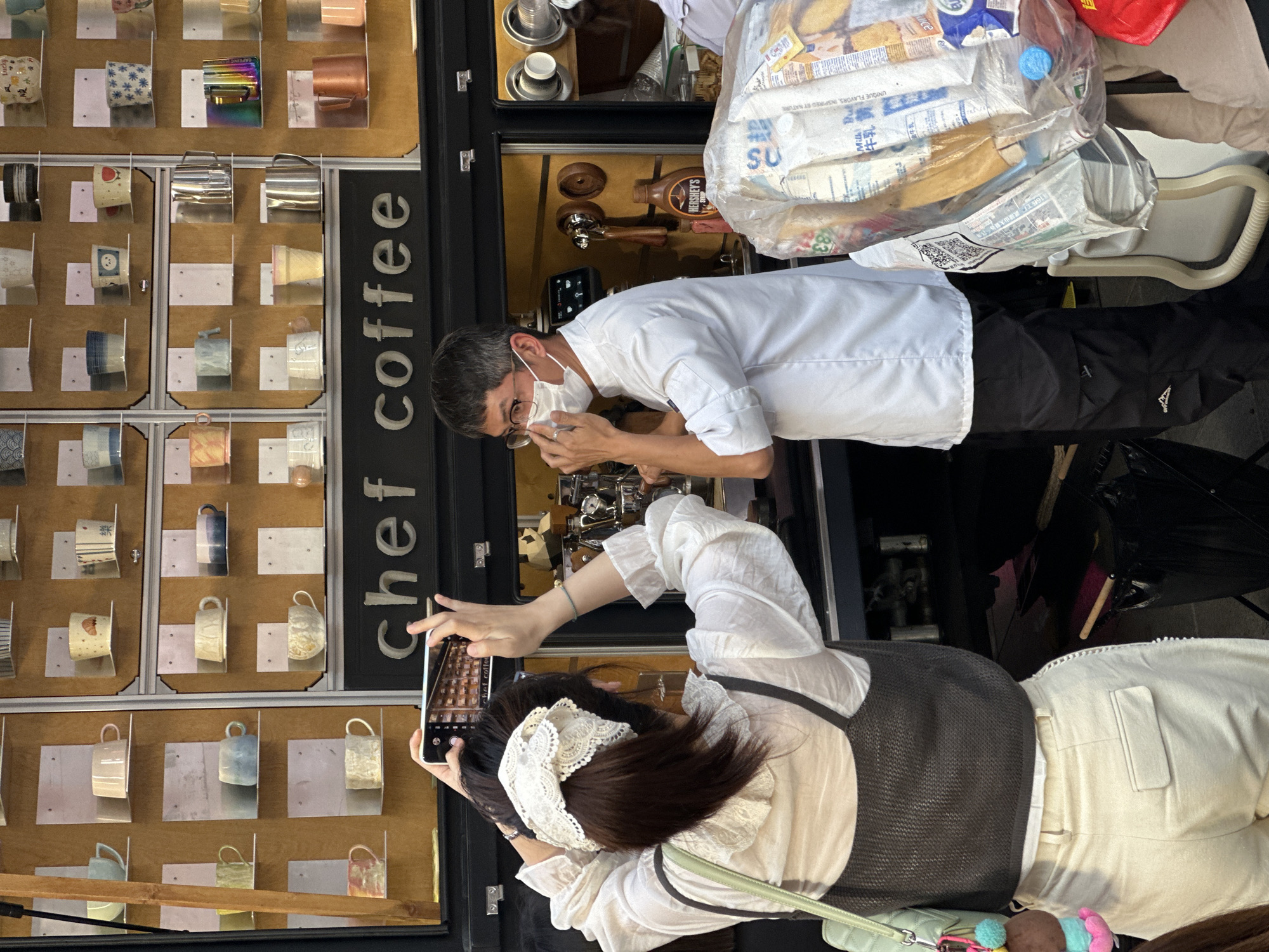 This daytime image captures a vibrant scene at an outdoor or semi-outdoor coffee stand in Yuzhong District, China. A male barista, wearing a white chef's coat and a face mask, is diligently preparing coffee at an espresso machine. He is leaning forward, hands near the portafilter, with a bottle of Hershey's chocolate syrup visible behind him.
To his left, a woman, viewed from behind, is holding a smartphone and appears to be taking a photo or video. She wears a white ruffled blouse, a dark sleeveless top, light-colored pants, and a white lace bow in her dark hair. A light green handbag with a colorful charm hangs from her shoulder.
The setting includes a prominent display wall to the left, showcasing a variety of coffee mugs and cups in different colors and patterns, some bearing text like "Lucky" and the Chinese character "樂" (le, meaning joy). A vertical black sign clearly displays "chef coffee". Behind the barista, plastic bags filled with supplies, including milk cartons labeled "Milk" and "牛乳" (Chinese for milk), and other goods with text such as "UNIQUE FLAVORS, INSPIRED BY NATURE" and "16杯工厂 武陵山大家", suggest a busy operational environment. The bright lighting indicates fair weather.