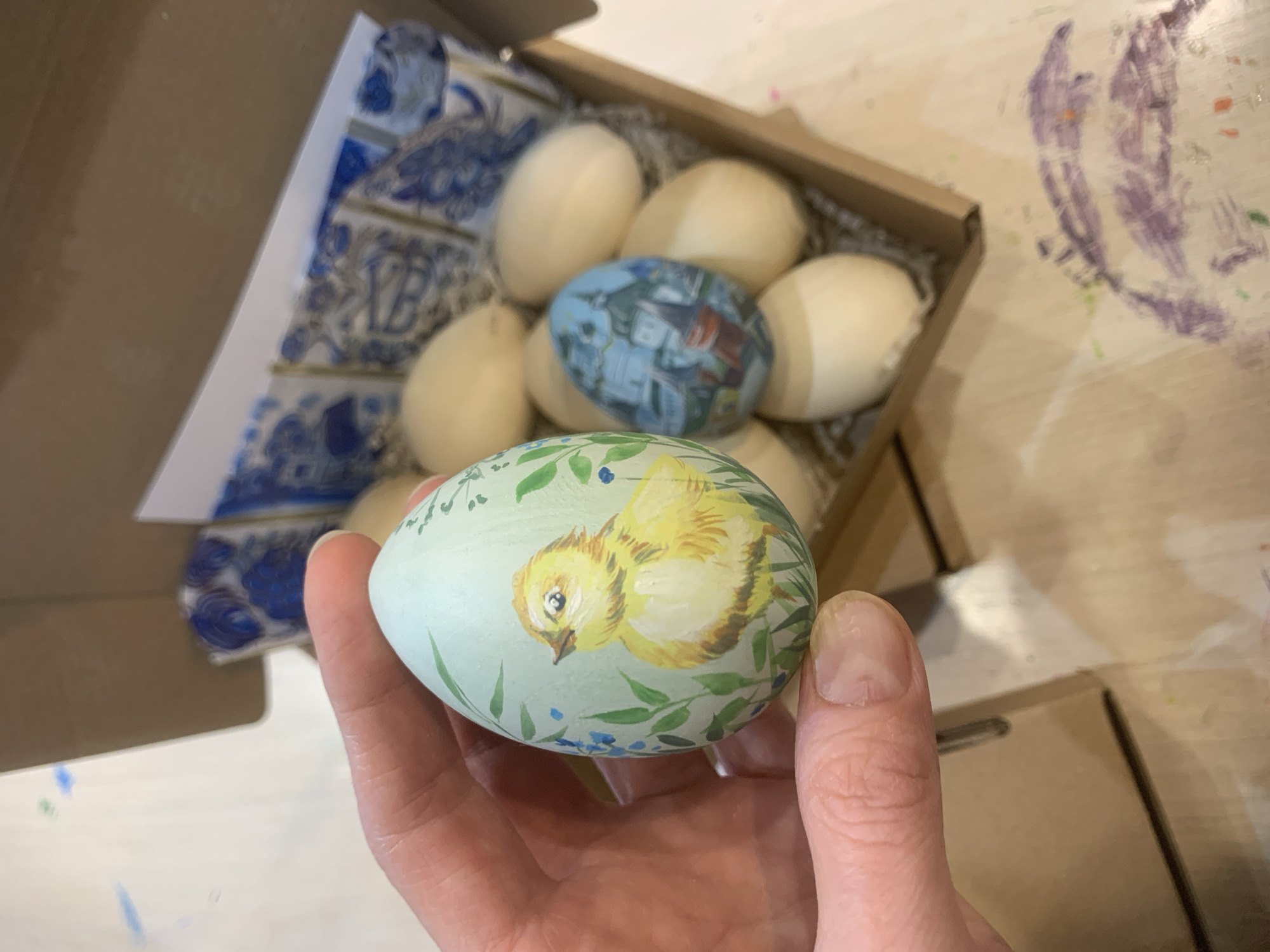 A person is holding a painted egg with a chick design in their hand. The egg is light blue with a yellow chick, green leaves, and small blue flowers. Behind the hand and egg is an open cardboard box filled with white and painted eggs nestled in shredded paper. Some of the eggs have blue designs. The box sits on a wooden surface with paint splatters visible. Based on the presence of a painted Easter egg and other eggs, this is probably a scene related to Easter. The location is not discernible from the image.