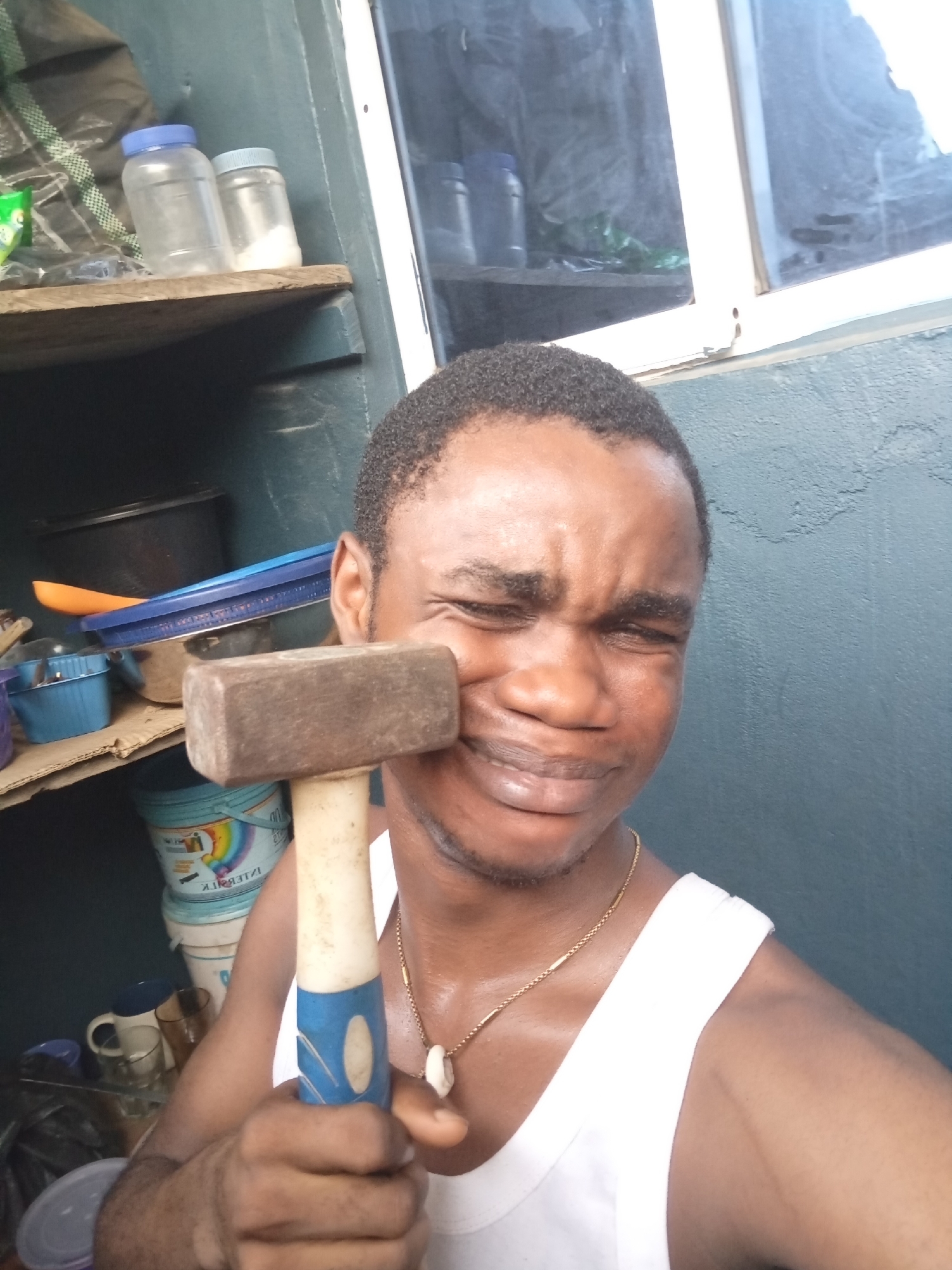 A man is holding a hammer against his face. He is wearing a white tank top and a gold chain. The background contains shelves with various containers, pots, and paint buckets. A window is visible in the upper right of the frame. The photograph appears to be taken indoors. The location is possibly in Owerri, Nigeria.