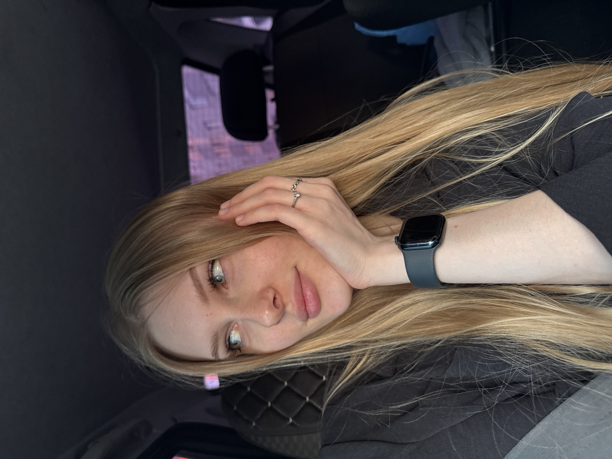 A fair-skinned, blonde woman rests her head on her hand, wearing a dark-colored smart watch and a silver ring on her right hand, likely sitting inside a vehicle, with the surroundings outside the window suggesting a location in Кондратово, Russia.