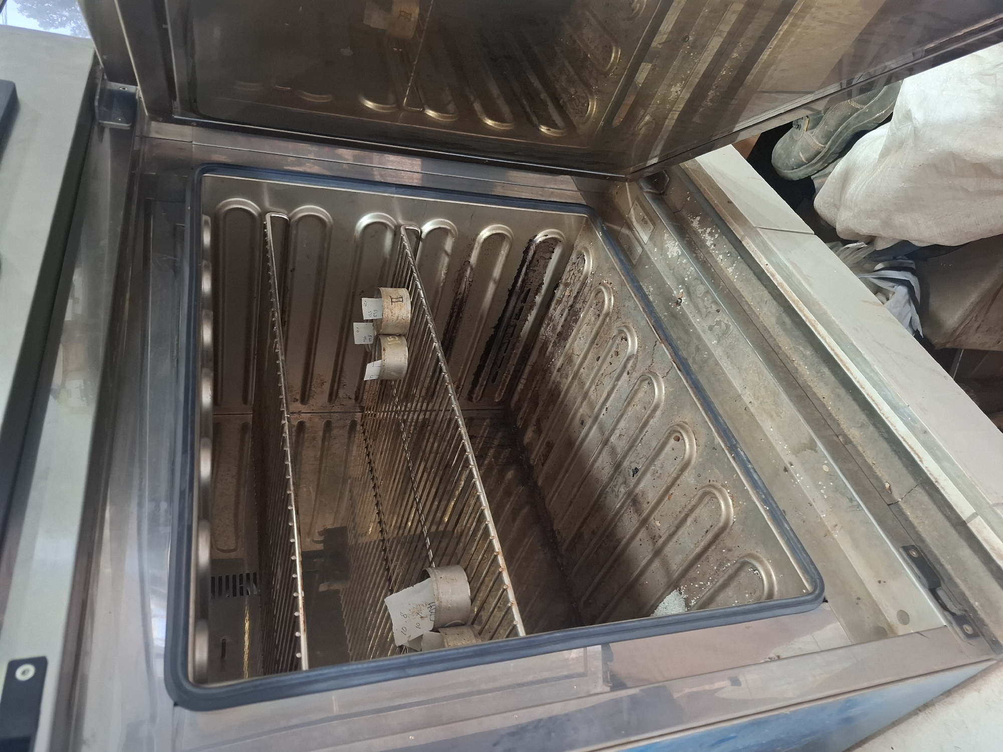 An open, commercial oven is shown. Inside the oven are several metal racks and what appear to be small metal containers with paper labels. The oven appears to be dirty, with residue on the racks and the bottom of the oven. Outside the oven, a shoe and some fabric are visible. The location is identified as Ciheulang Tonggoh, Indonesia.