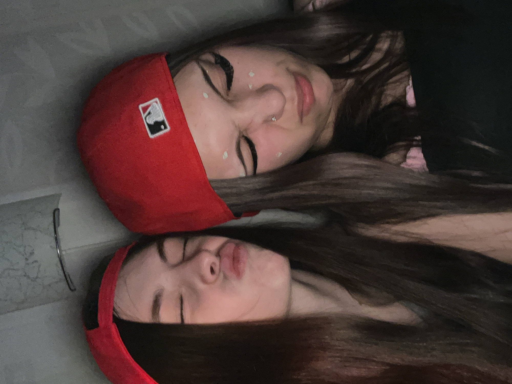 Two young women are captured in a close-up, top-down shot, lying side-by-side with their heads close together. Both have long, dark hair spread out and are wearing red baseball caps.
The woman positioned higher in the image (to the right in the original horizontal photo orientation) has her eyes squinted or closed and a soft smile. She has a small nose piercing and several distinct white dots, possibly makeup or decorative elements, on her cheeks and forehead. Her red baseball cap features a clear white MLB (Major League Baseball) logo on its front panel.
The woman positioned lower in the image (to the left in the original horizontal photo orientation) is making a playful "pouty" face with her lips pursed, and her eyes are closed. She also appears to have similar white dots on her face.
The background is a simple, light gray surface, possibly a bed, floor, or couch, suggesting an indoor setting. A thin, metallic object, resembling glasses or a hair clip, lies on the gray surface near the lower woman's cap. The lighting is dim, contributing to a relaxed and intimate atmosphere. The overall activity appears to be a casual, playful moment captured in a selfie or close-up photo.