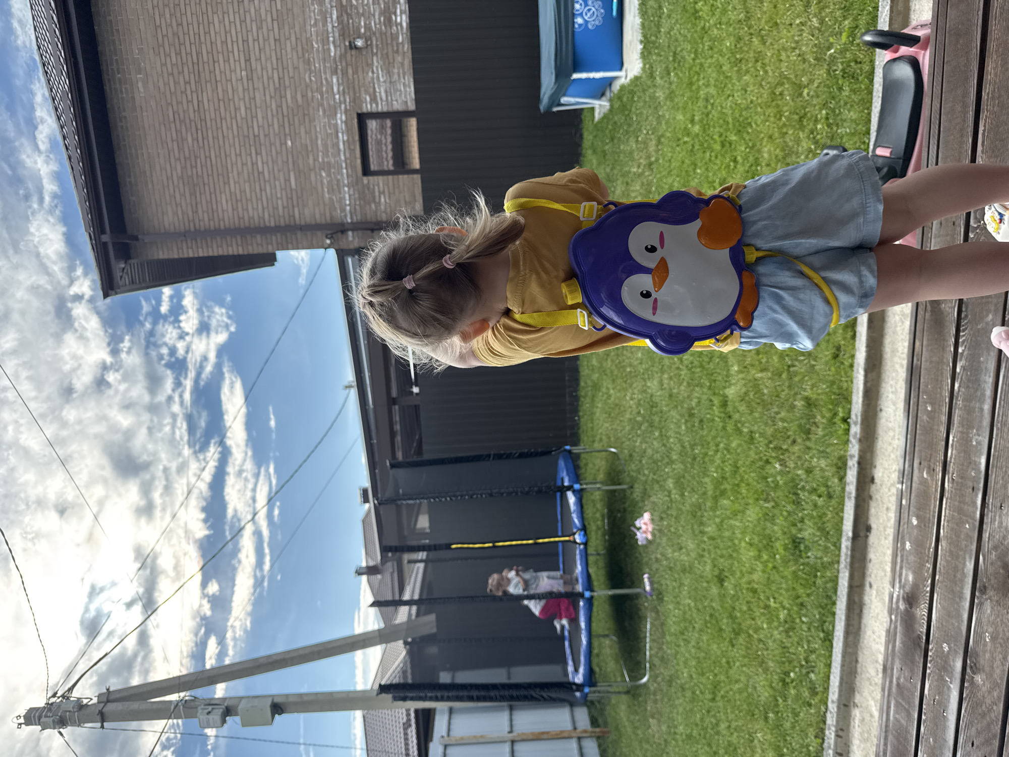 In an outdoor backyard setting, likely in the city of Дёмино, Russia, a young girl faces away from the camera, wearing a yellow t-shirt, light blue shorts, and a purple penguin-shaped water backpack. She has blonde hair styled in two pigtails. In the background, another child is visible on a trampoline. The backyard features a trampoline with a safety net, a small blue kiddie pool, a wooden fence, and a tall utility pole with electrical wires. The ground is covered in green grass. A pink scooter is partially visible in the foreground. The sky is partly cloudy.