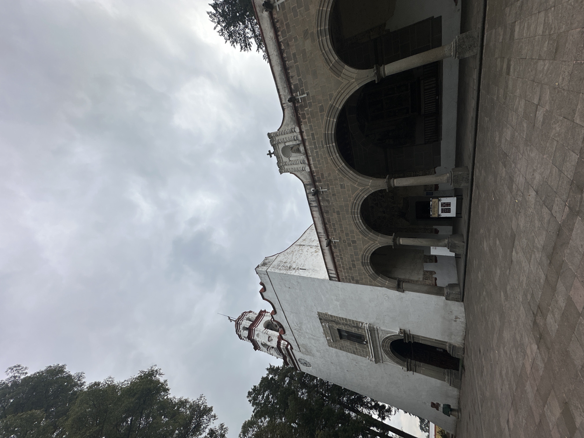 The image shows the Ex Convento del Santo Nombre de Jesús, located in San Miguel Zinacantepec, Mexico. The architecture features a combination of white walls with red trim and gray stone. The building has a tower and a covered arcade supported by columns. The sky is overcast with gray clouds. Trees are visible at the bottom of the frame. A brick or stone-paved walkway leads to the building. No people are clearly visible.