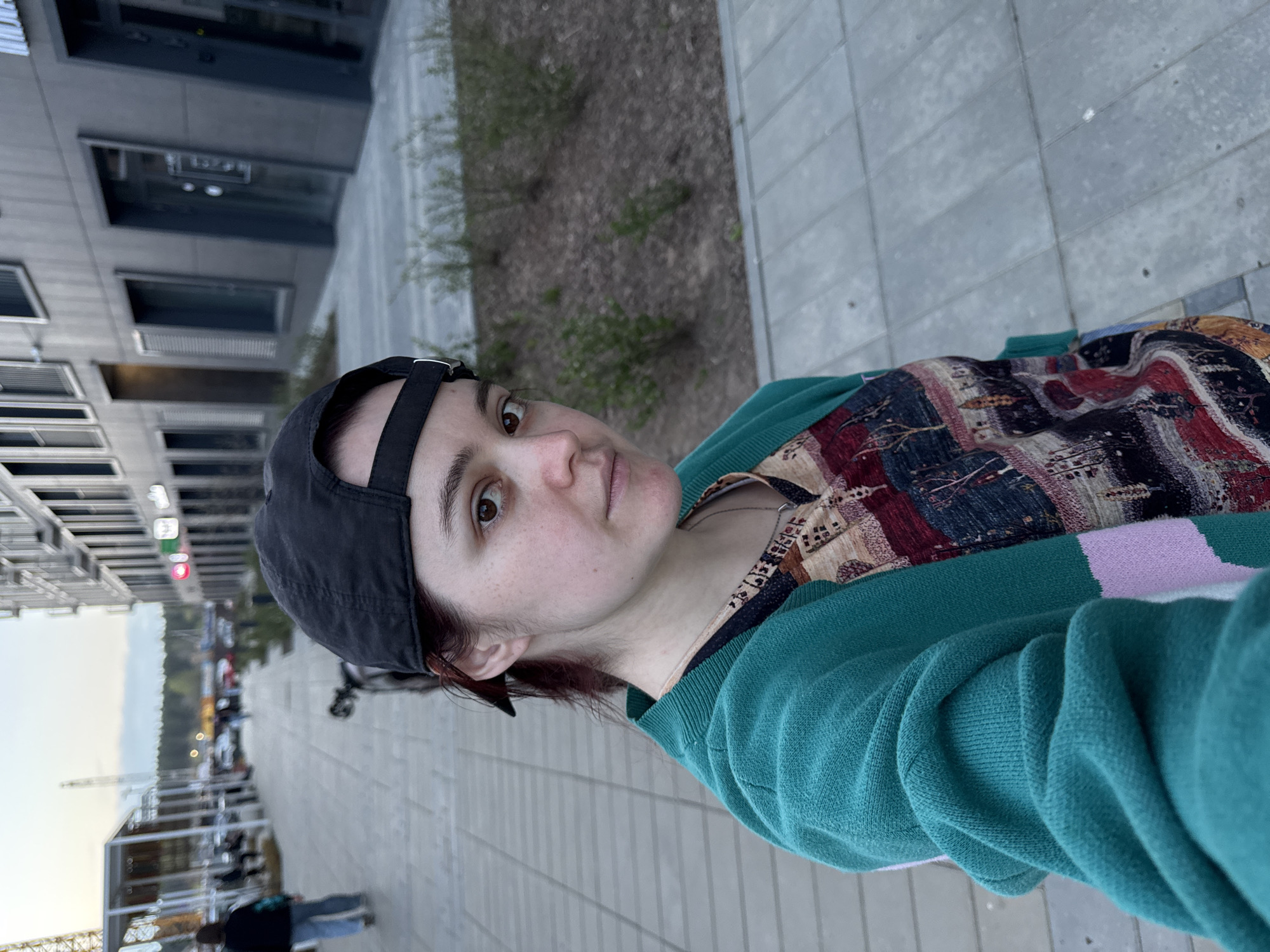 A person with fair skin is seen from the chest up, angled slightly towards the right, against an urban backdrop. The person wears a black baseball cap, a patterned top featuring burgundy, beige, and black designs, and a teal-colored sweater with a light purple stripe. The background includes a building with multiple windows, a pedestrian walkway paved with grey rectangular stones, and a patch of ground with vegetation. In the distant background, city infrastructure is visible. Based on the location tags and landmarks, the location is Izhevsk, Russia.