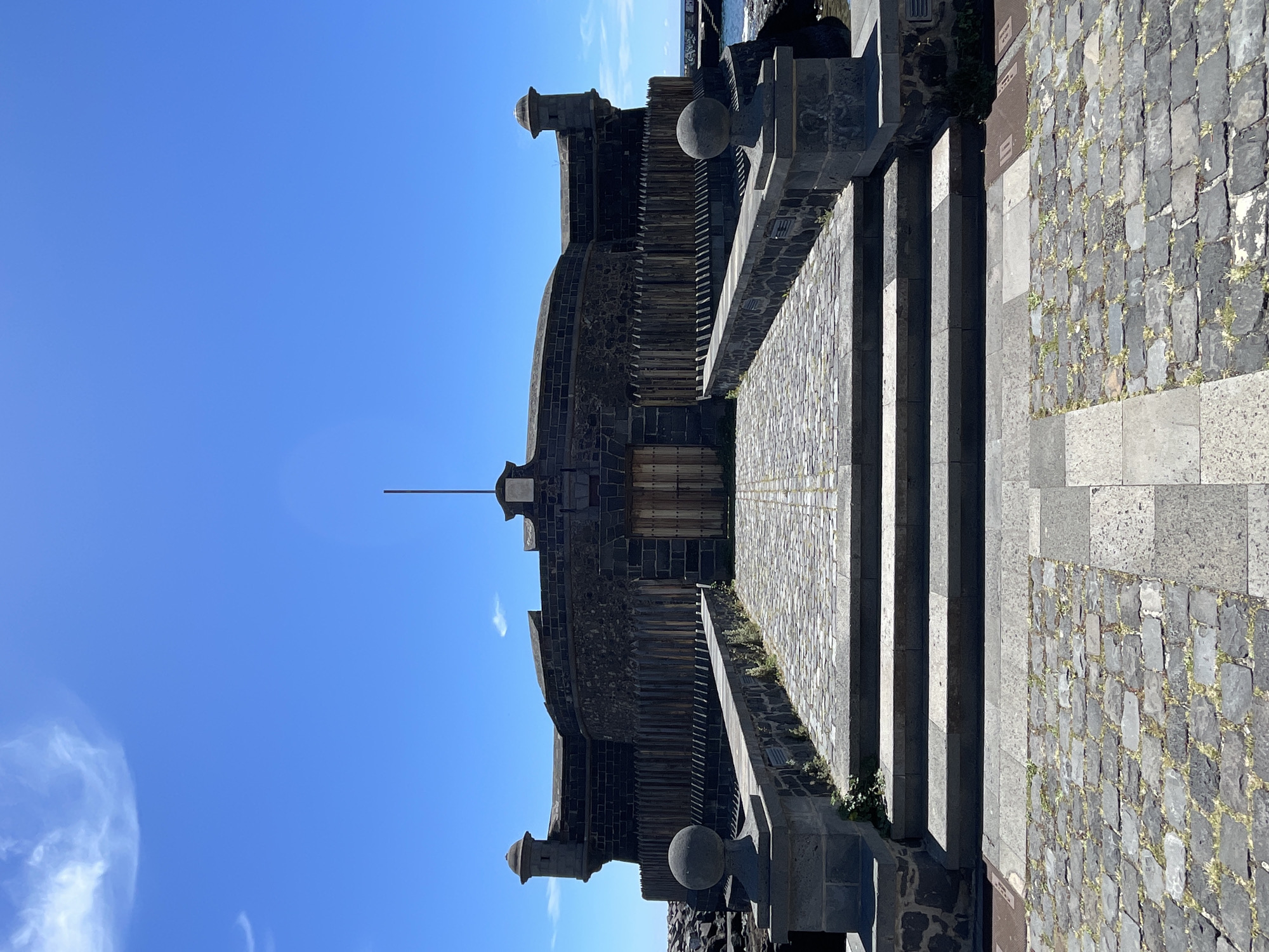 The image shows the Castillo de San Juan Bautista, also known as the Black Castle, in Santa Cruz de Tenerife, Spain. It is oriented vertically with the castle in the center. The fort is constructed of dark stone and has a simple, fortress-like design with a central body and two cylindrical turrets with domes on either side. In front of the castle, there is a stone pathway with steps that lead to the entrance. The path is made of cobblestones and stone slabs. The sky is clear and blue, with a few wispy clouds visible. The ocean is visible in the top right corner of the image. The structure appears to be well-maintained, and no people are visible in the scene.