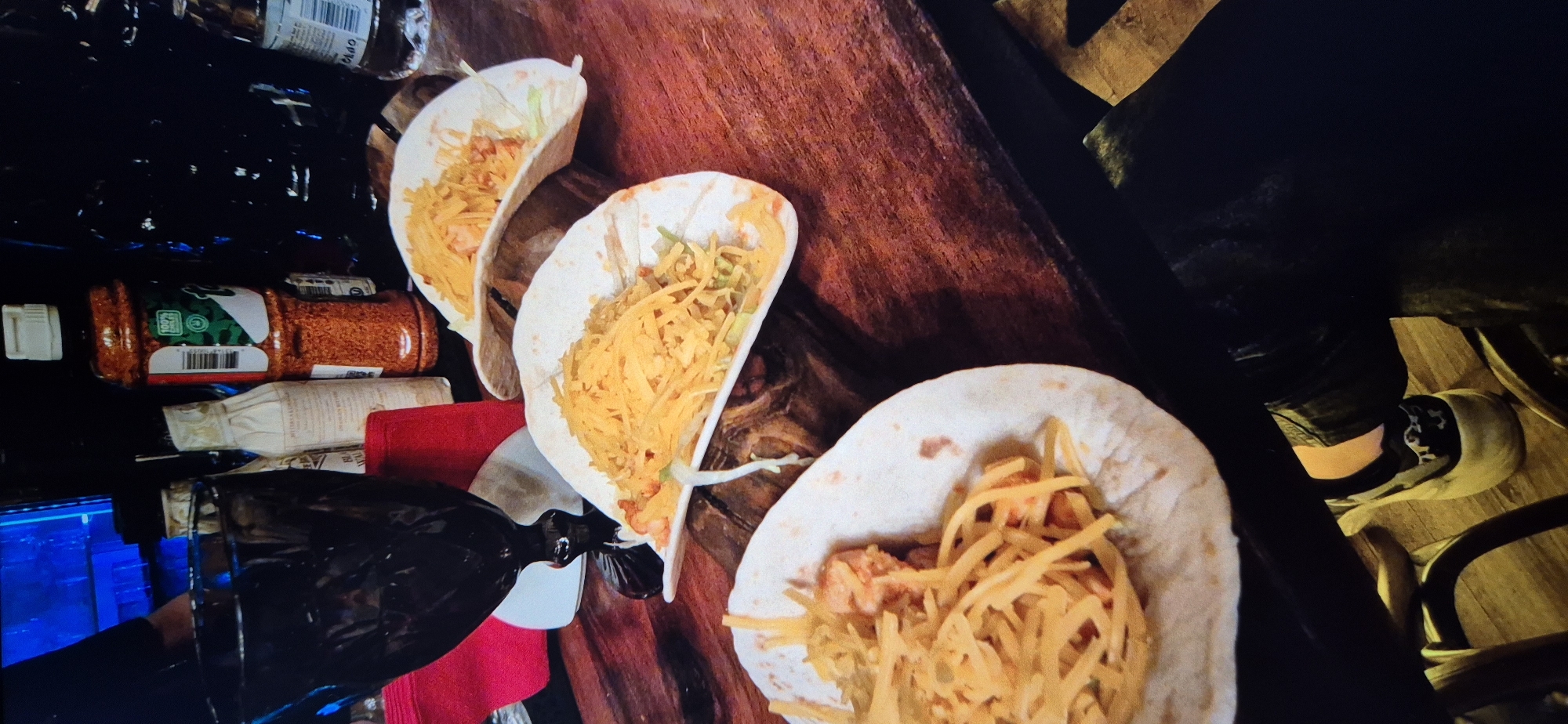 The image depicts a countertop cluttered with items and three tacos. The tacos are filled with shredded cheese and lettuce. Adjacent to the tacos are several bottles, including a bottle of what appears to be chili flakes and a few bottles of liquids, arranged somewhat haphazardly. A black object partially obscures the view of some of the items on the counter. In the background to the right, a leg and foot wearing a shoe are visible. There is no evidence to ascertain the location is in Sofia, Bulgaria.