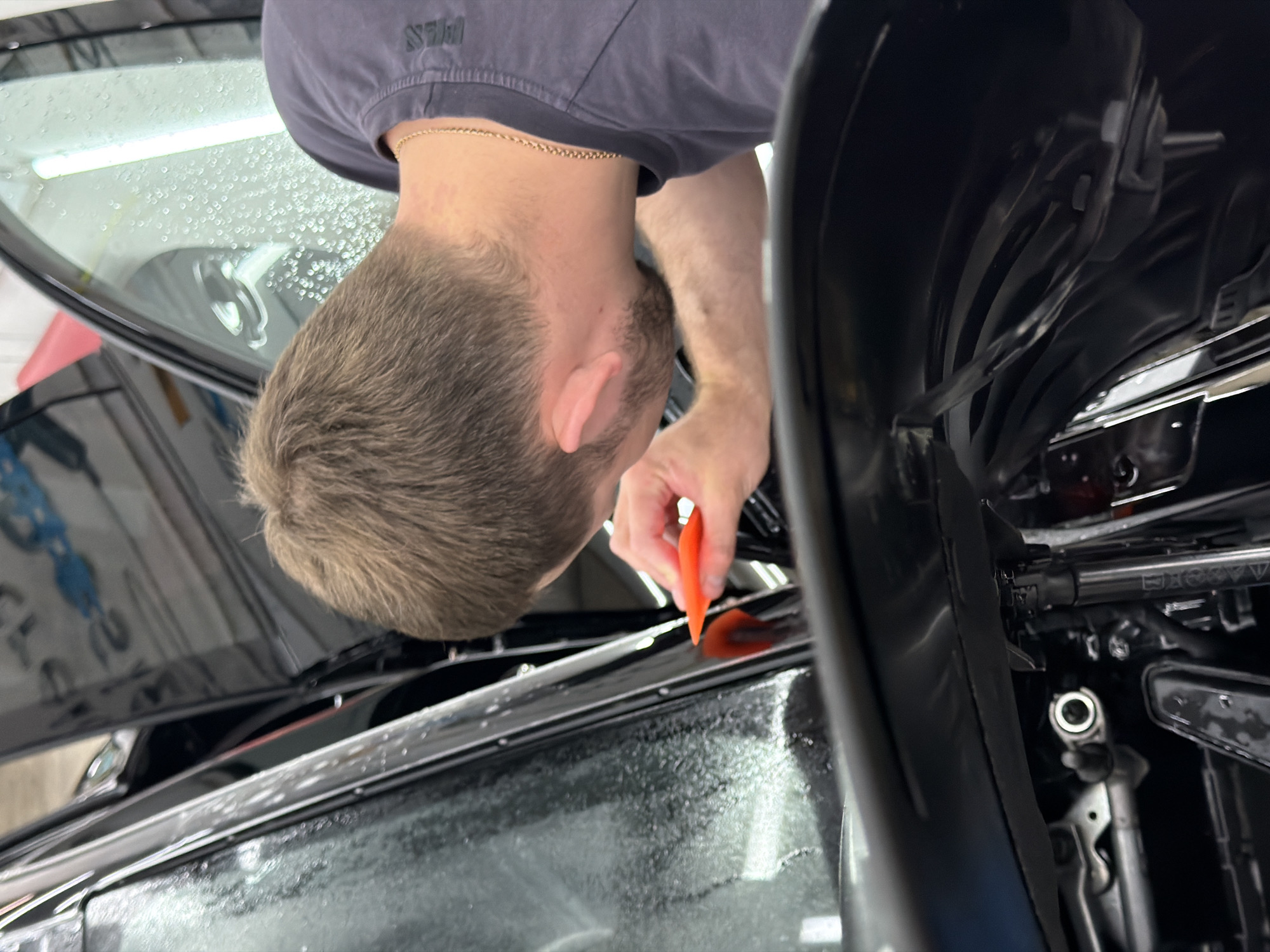 A man is using an orange tool to apply film to a car window. The man has short brown hair and is wearing a gray t-shirt and gold chain. The car is black and appears to be inside a garage. The exact location in Krasnoyarsk, Russia is not evident from the image.