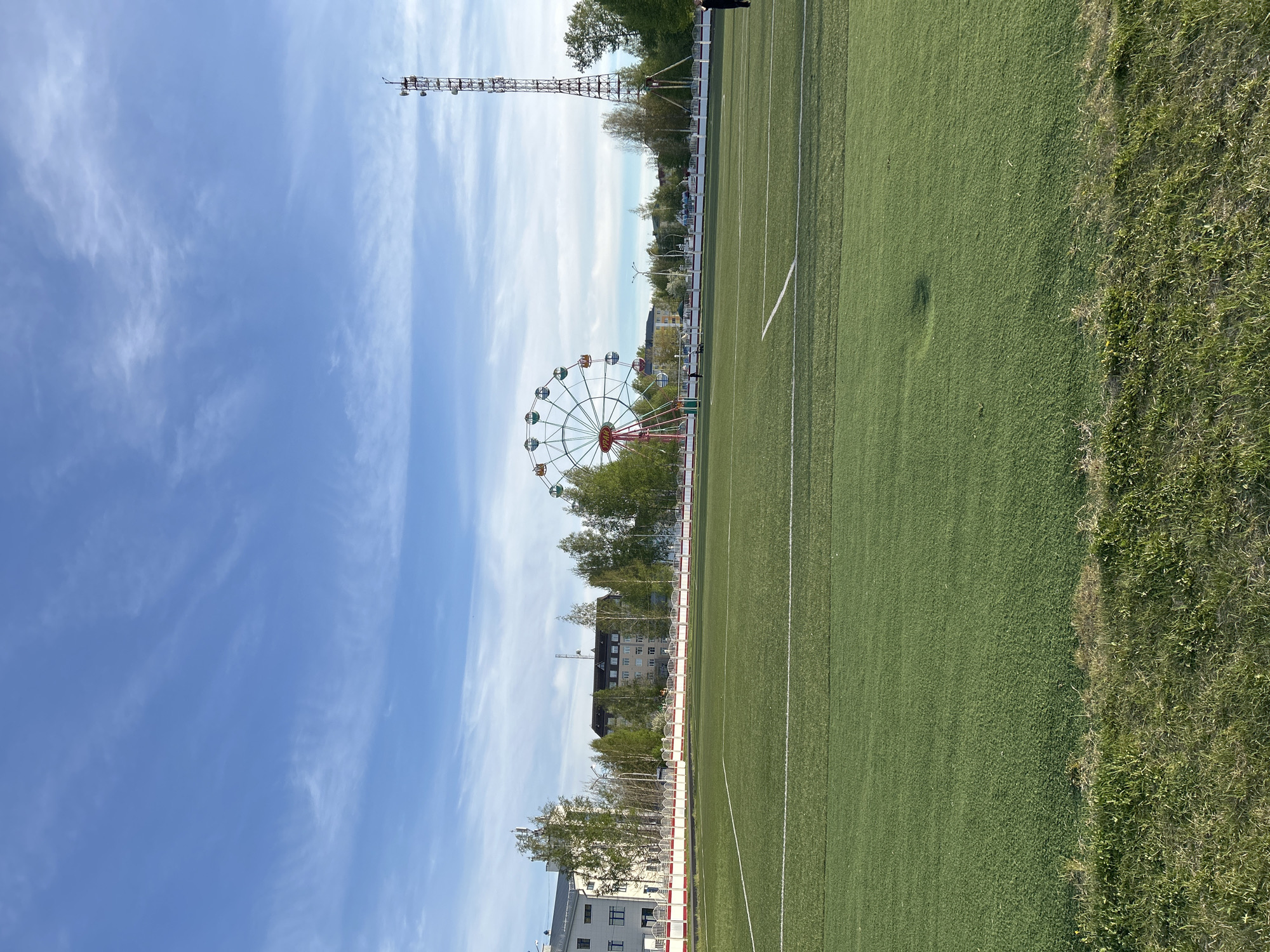 The image depicts a view of the Uray Stadium in Uray, Russia. A Ferris wheel is visible in the background, positioned beyond the stadium's perimeter. The stadium itself has a green artificial turf field with white lines marking its boundaries. A tall transmission tower stands near the stadium, extending high into the blue sky with scattered clouds. The foreground shows the green sports field, transitioning into a rougher grassy area on the right side of the frame. Buildings can be seen amongst the trees beyond the stadium fence.