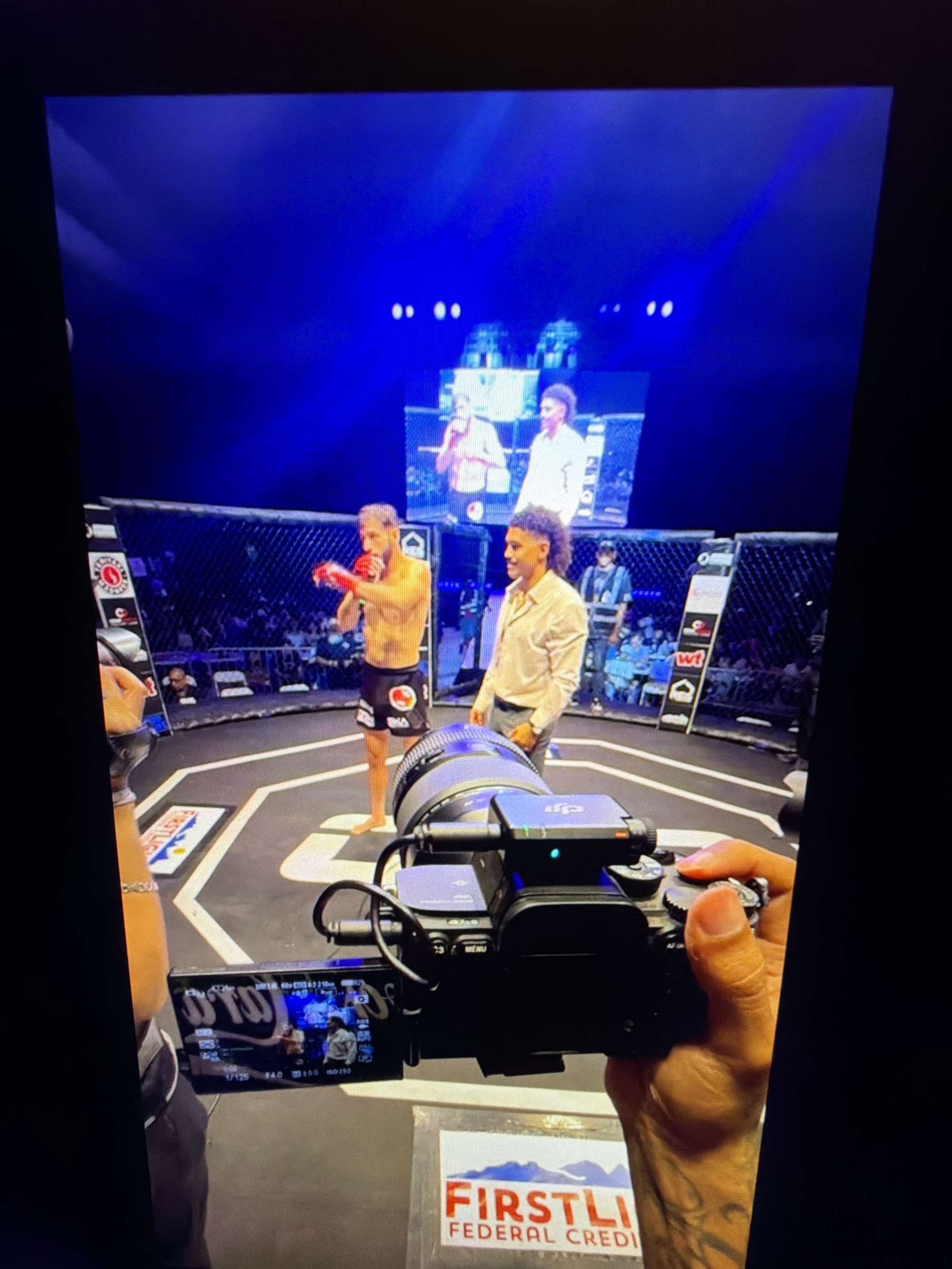 The image shows a boxing ring with two male fighters in the center, both wearing boxing gloves and shorts. One fighter, on the left, is wearing black shorts with a logo and is in a defensive stance. The other fighter, on the right, is wearing light-colored shorts and is looking towards the camera. In the background, a large screen displays images of people. A cameraman is in the foreground, holding a professional camera. The scene appears to be a live event, possibly a boxing match or competition. The environment suggests an indoor venue with a spotlighted ring and a crowd in the background. The presence of a cameraman and a screen indicates a broadcast or recording of the event. The image does not contain any specific details that would allow for the identification of the city of Kafr Yasif, Israel.