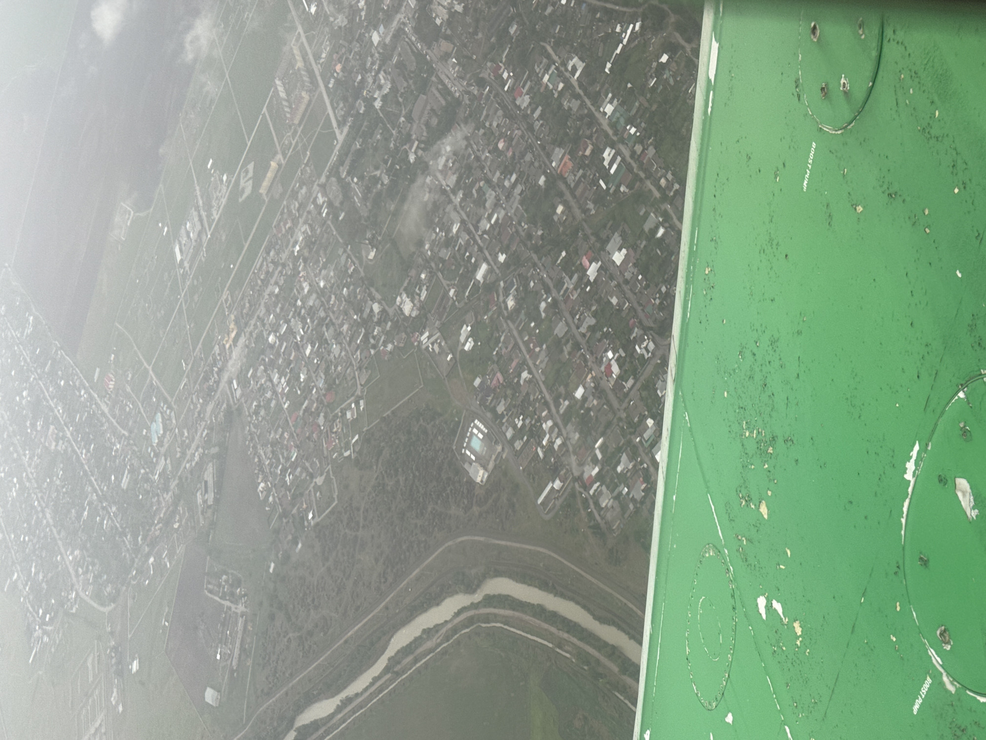 The image depicts an aerial view of a town in Bishkek, Kyrgyzstan, taken from inside a plane. The right side of the frame shows part of the plane's interior, specifically a green panel with visible wear and labels such as "BOOST PUMP". The landscape below consists of a dense residential area with numerous buildings and houses, intersected by roads and pathways. A river meanders through the lower-middle ground of the image. To the left, the scene is obscured by what appears to be fog or haze, reducing visibility.