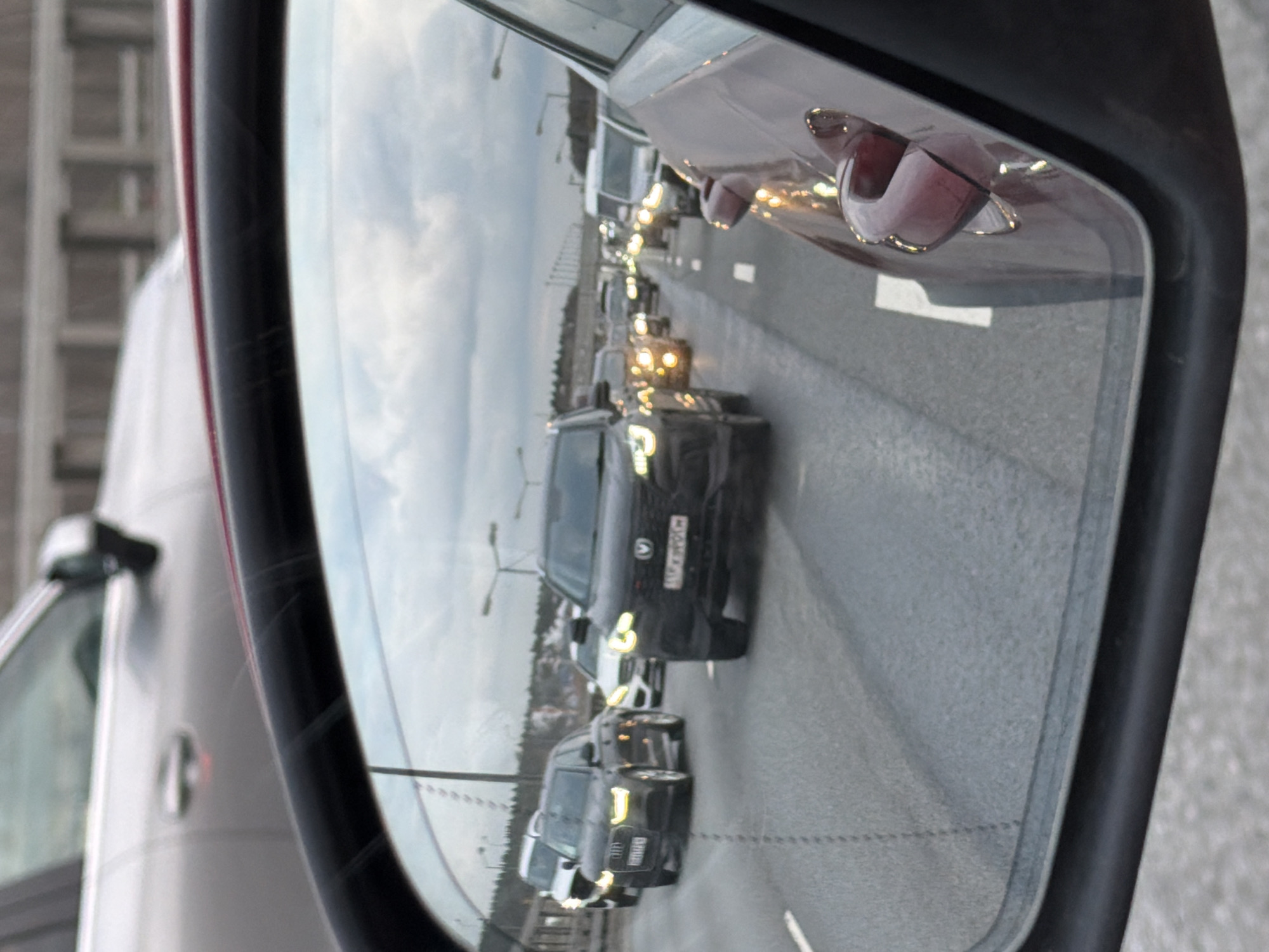The image displays a view through a car's side-view mirror, showing a traffic jam on a multi-lane road under an overcast sky. The perspective is from inside a vehicle, with parts of its own body visible: a red door handle and a section of a maroon car door on the right of the mirror, and a blurred grey interior or body part on the left.
Within the mirror's reflection, numerous cars, mostly dark-colored sedans and SUVs, are visible in a queue, indicating slow-moving or stopped traffic. The most prominent vehicle in the reflection is a black SUV, on which a Russian license plate "B020XA 799" is clearly visible. The "799" region code confirms the location context of Moscow, Russia. Further along the road, other vehicles have their headlights or daytime running lights illuminated, creating bright points of light against the grey road surface. Streetlights are also visible in the distance. The road itself features white lane markings.
The time of day appears to be daytime, possibly late afternoon, given the cloudy sky and the illuminated car lights. The overall scene captures a typical urban traffic congestion event.