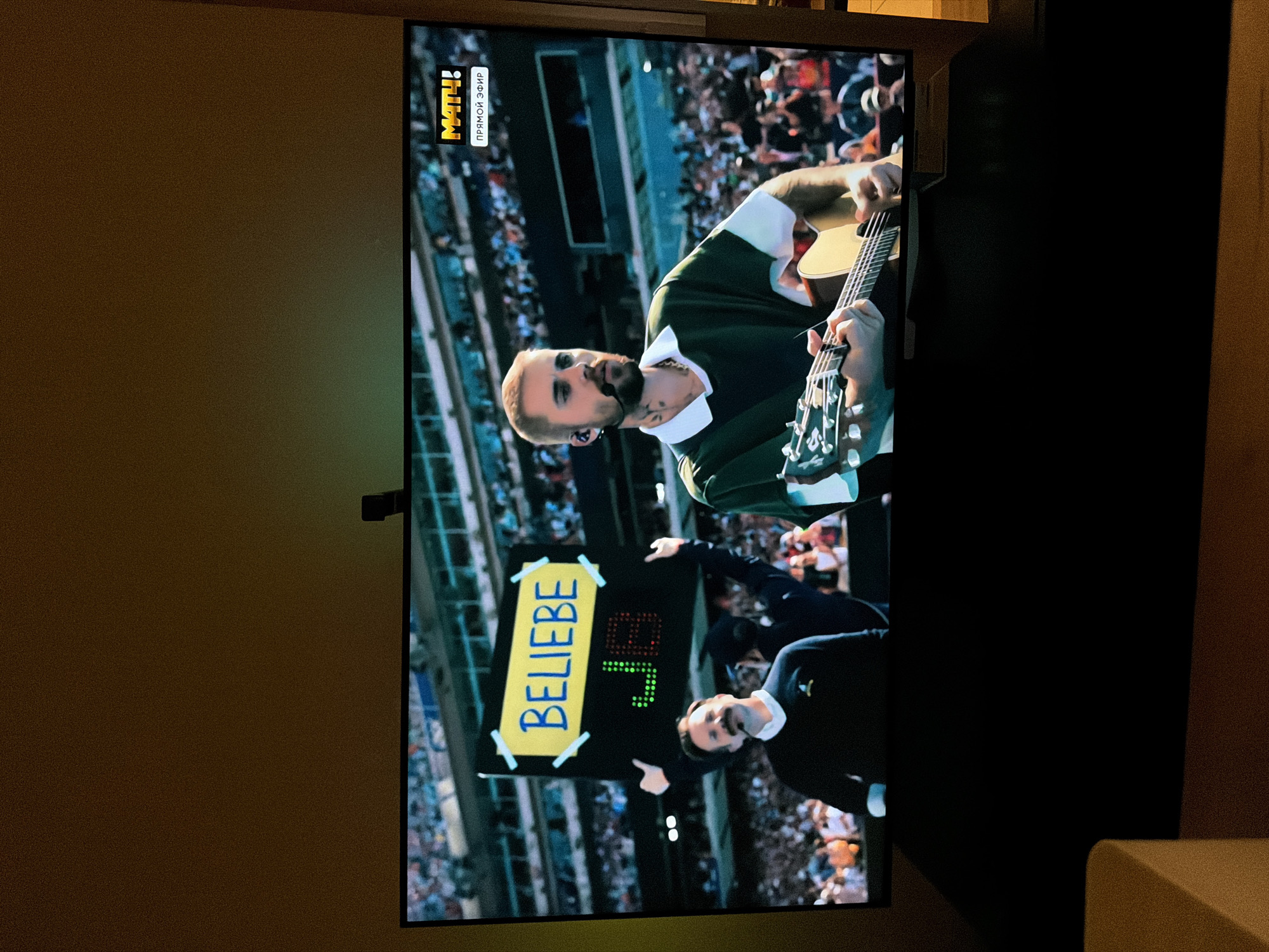 The image shows a screen displaying a live broadcast, likely from a sporting event given the "МАТЧ!" logo in the upper right corner, which translates to "MATCH!". The broadcast features a male musician playing an acoustic guitar on a stage with a large audience in the background, seated in stadium bleachers. In the foreground on the left side of the screen, a man with a mustache and wearing a dark sweater is looking towards the camera. Behind him, another person is holding up a sign that says "BELIEBE JB" in blue lettering on a yellow background, with "JB" displayed in a green and red LED matrix. The overall scene depicts an entertainment performance within a large venue. The location is specified as the city of Khimki, Russia.