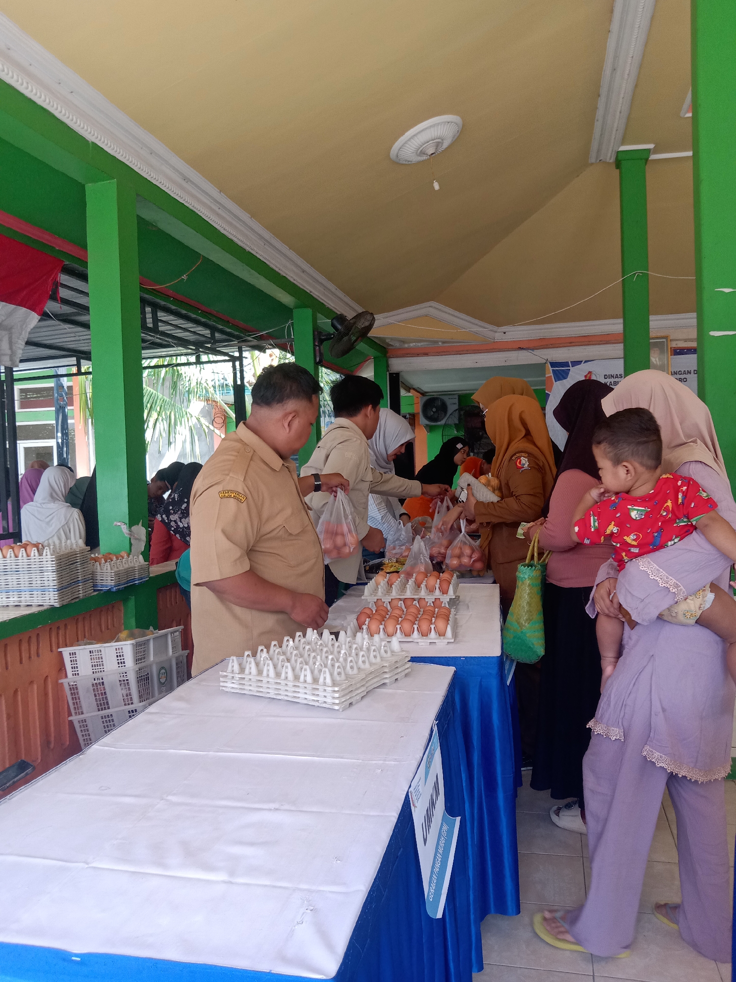 The image depicts an outdoor market scene in Klepek, Indonesia, possibly at a community event or distribution point. In the foreground, a man in a tan uniform, likely a government official, is holding a plastic bag of eggs. He stands behind a table covered with a white cloth, on which several trays of eggs are displayed. To the right, a woman in a lilac outfit is carrying a young child on her hip. She is looking at the food items being distributed. Other people, mostly women wearing hijabs, are visible in the background, interacting with each other and with what appear to be food vendors or organizers. The environment is characterized by green pillars, a yellow ceiling, and a tiled floor. Banners and signs are partially visible, suggesting an official event. The overall atmosphere appears to be one of community gathering and distribution of essential goods.