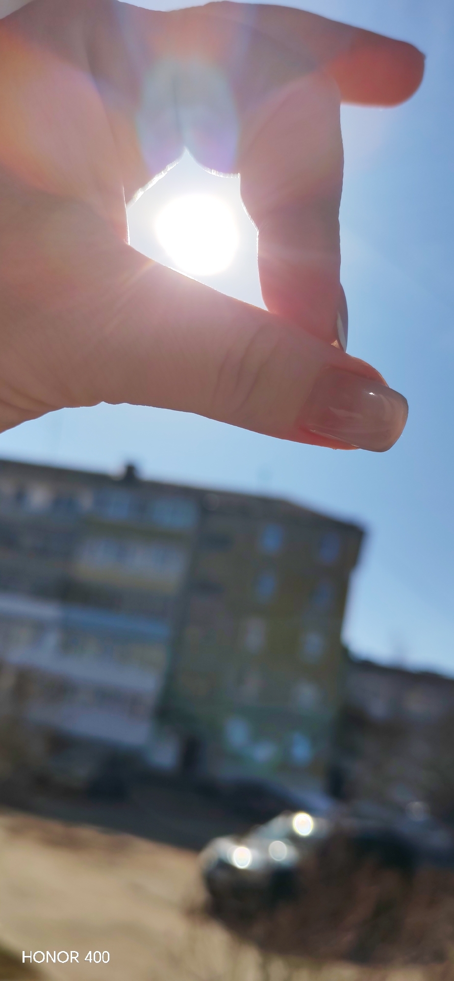 A hand is holding the sun, partially obscuring the light with the tips of the thumb and forefinger, against a blue sky. In the background, out of focus, is an apartment building and a car. The image was taken with an HONOR 400. The exact location in Noviy, Russia, cannot be determined from the image.