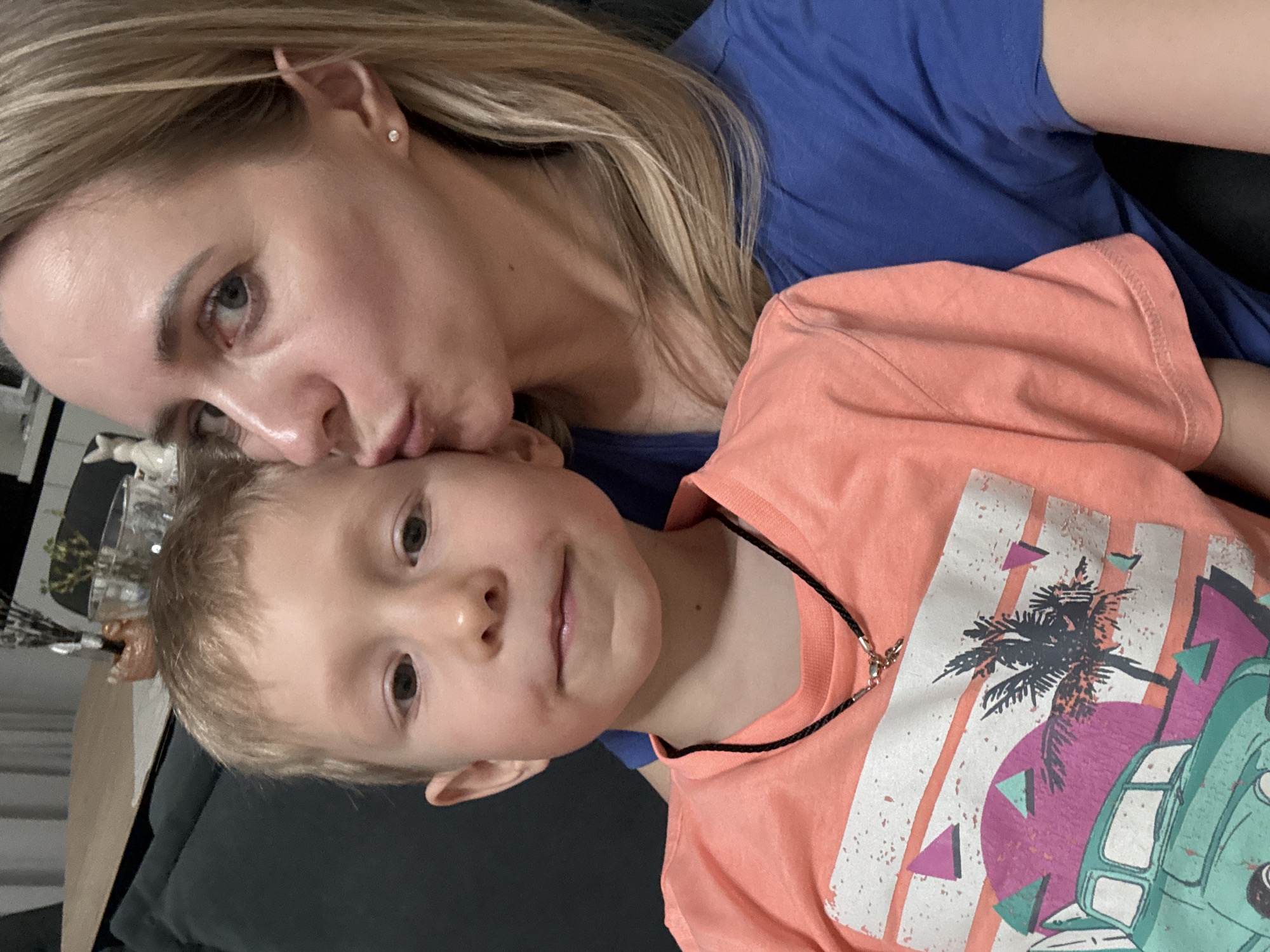 A woman with blonde hair is kissing the forehead of a young boy wearing a coral-colored t-shirt with a palm tree and van design. The woman is wearing a blue shirt. They are likely inside, possibly sitting on a dark-colored couch. The location, Chaikovsky, Russia, cannot be independently verified from the image alone.