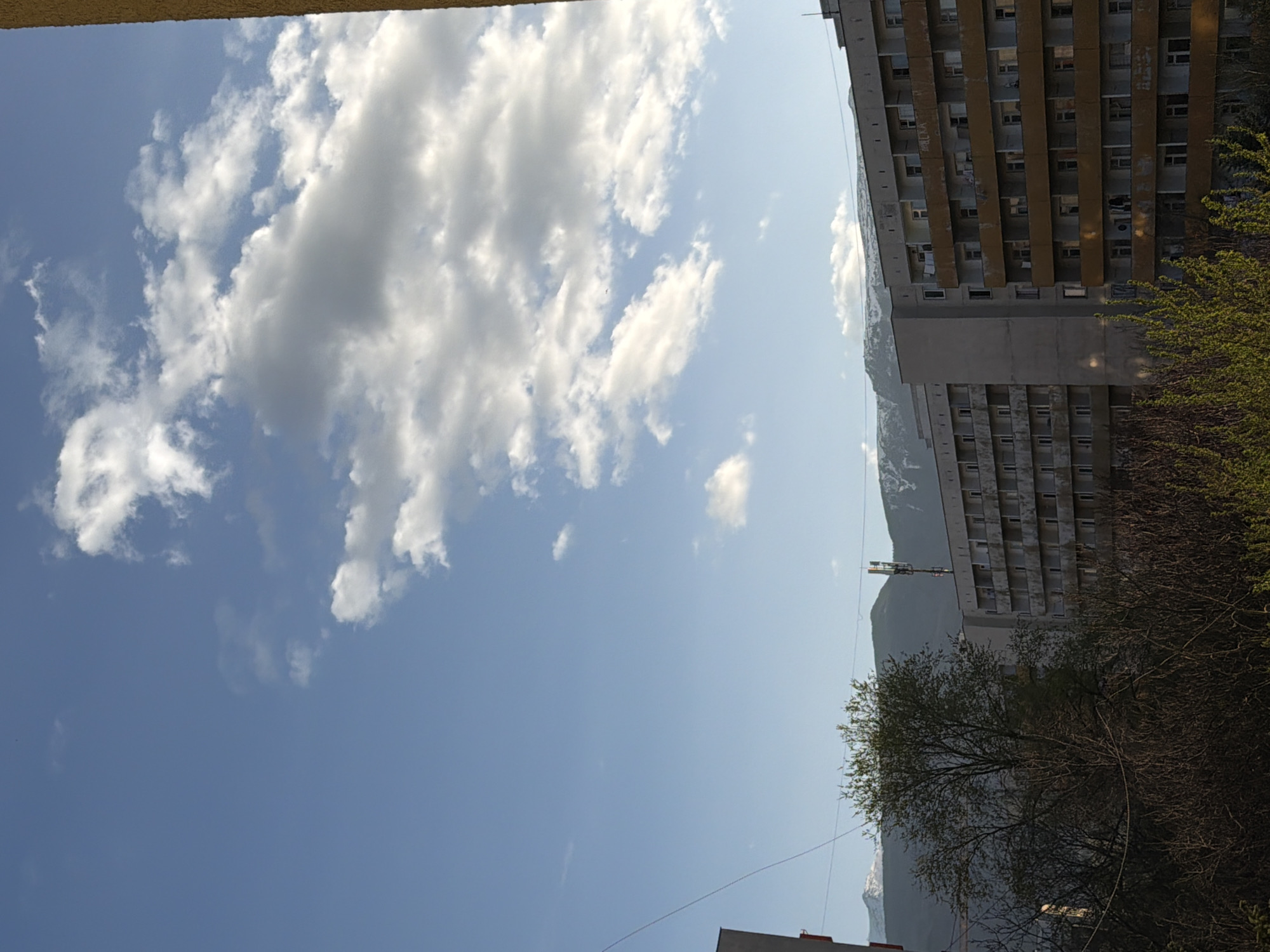 The image presents a view of the sky with numerous clouds in the left half, transitioning to a clear blue sky in the lower portion. A tall building with multiple windows and balconies is on the right side of the frame. Part of a mountain with snow can be seen behind the building. Trees are visible near the building's right side. Electrical wires are present in the lower part of the image. The location is likely Sofia, Bulgaria, based on the architecture style and setting.