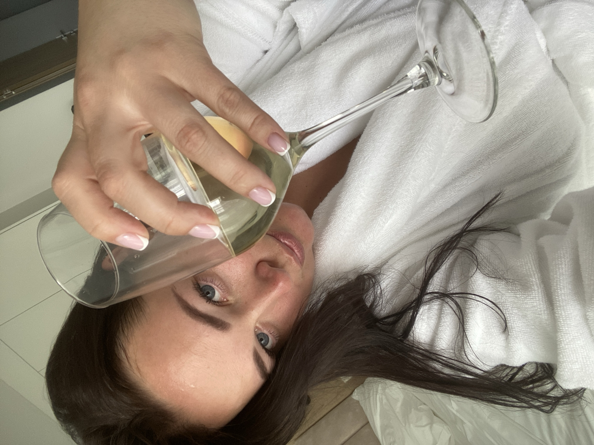 A woman is reclining, dressed in a white bathrobe, holding a wine glass filled with a light-colored liquid. Her brown hair is spread around her on the white surface. The location cannot be determined from the provided image.