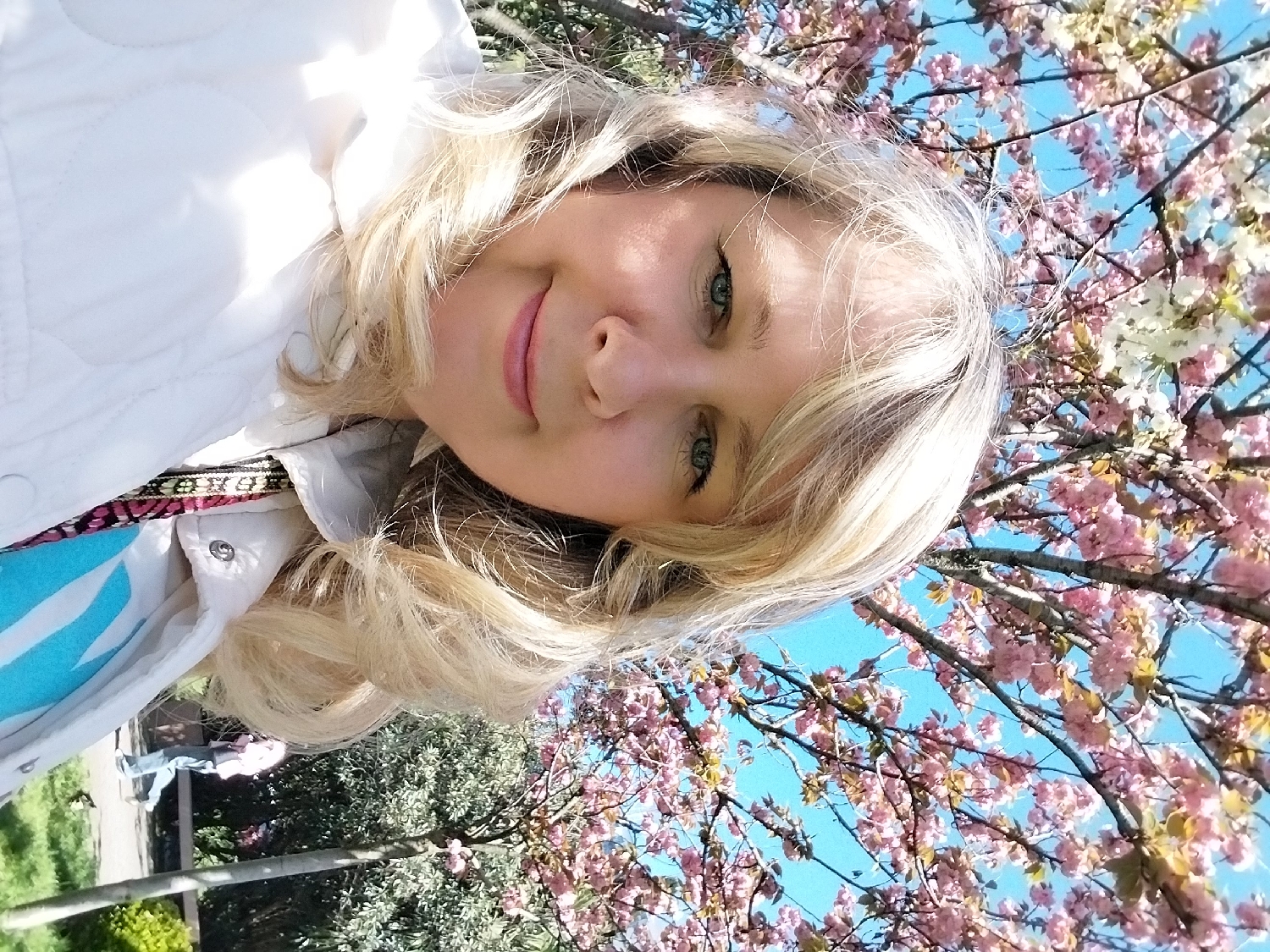 A blonde woman is featured in a close-up, rotated portrait, likely a selfie taken outdoors. She has light skin, light blue eyes, and is looking towards the camera with a subtle smile. Her long, wavy blonde hair frames her face. She is wearing a white quilted jacket over a light blue and white patterned garment, with a colorful patterned strap visible below her jacket. Bright sunlight is visible in the upper left of the frame.
The background is dominated by branches of trees laden with vibrant pink and some white blossoms against a clear, bright blue sky, suggesting springtime. The perspective is from a low angle, looking upwards. In the distance, faint figures of people are visible amidst green foliage and tree trunks, indicating a public outdoor setting such as a park or garden. The day appears sunny and pleasant. No text is visible in the image. The scene takes place in Sochi, Russia.