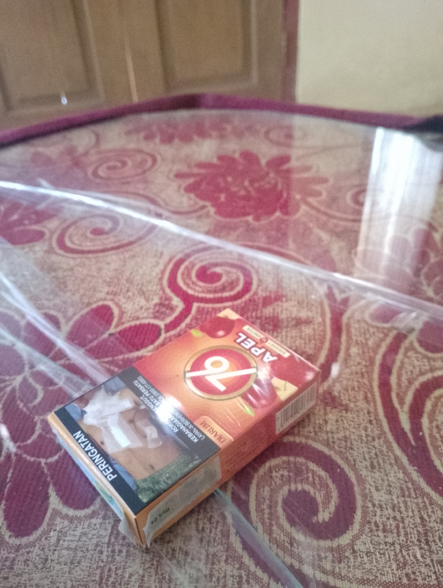 A pack of "Apel 76" cigarettes, a Indonesian brand, lies on a red patterned tablecloth. The pack is partially covered by a clear plastic sheet. The cigarettes are filter-tipped and the pack features warnings about the dangers of smoking. The background shows a wooden surface, possibly a table or wall. The scene is likely indoors, given the tablecloth and the general ambiance. The image does not provide any specific information to determine its exact location within Sukamanah, Indonesia.