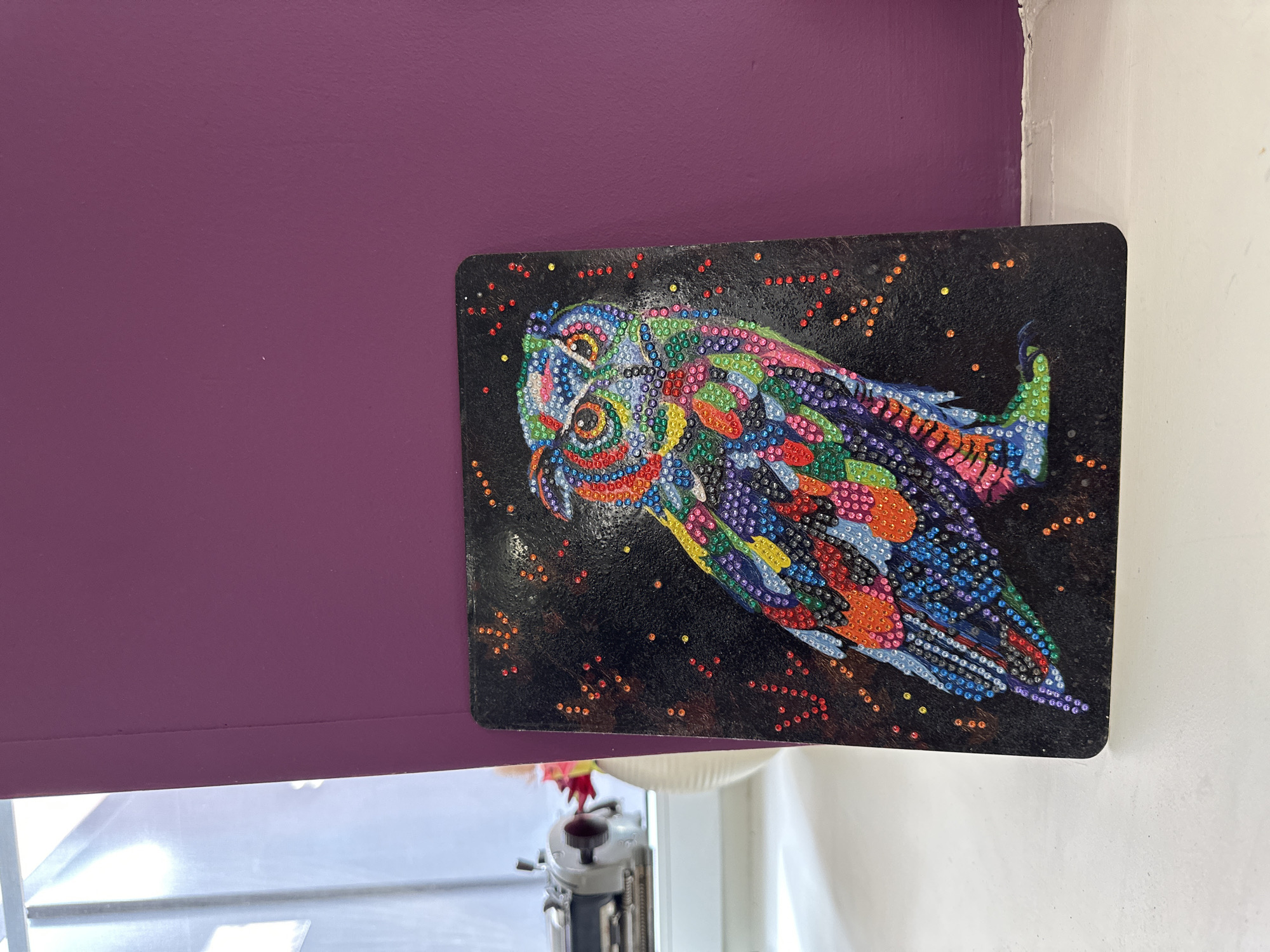 A painting of an owl is displayed against a purple wall in Saint Petersburg, Russia. The owl is brightly colored with reds, greens, blues, purples, and yellows on a black background with orange specks. The painting is rectangular with rounded edges and is mounted slightly askew on the wall. A white wall is visible to the right of the painting. Below the painting, part of a window blind and a white windowsill are visible. A sewing machine is visible through the partially opened window blind.