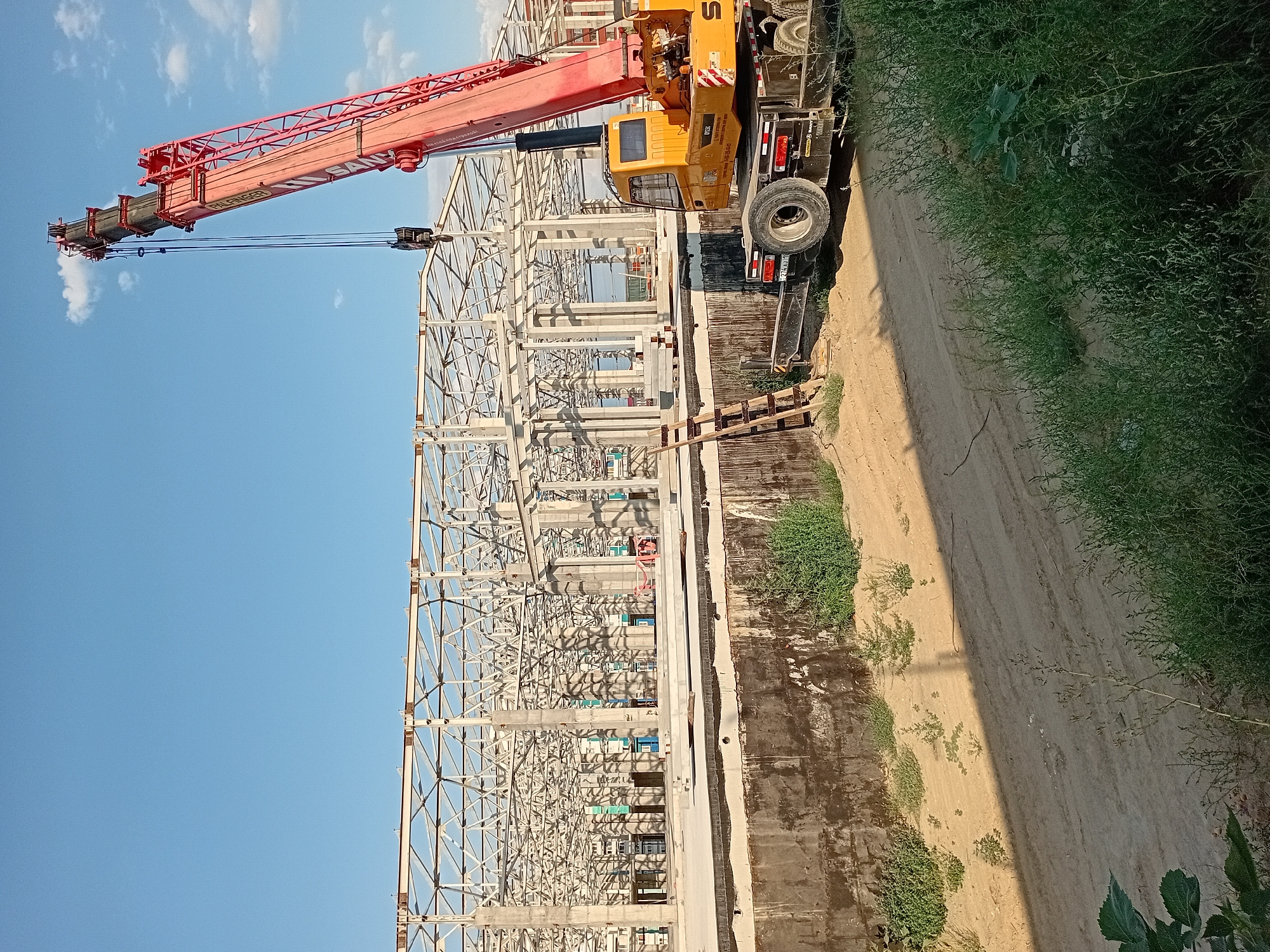 This image depicts a construction site in the city of Semey, Kazakhstan. A large, red mobile crane, with the brand "SANY" visible on its side, is positioned to the right of the frame. The crane's boom extends upwards and to the left, with a hook assembly suspended in the air, suggesting ongoing work. In the background, the skeletal structure of a large building, likely a warehouse or industrial facility, is under construction. The building features a complex network of white metal beams supported by concrete columns. The foreground shows a patch of dry, sandy ground with sparse green vegetation, indicating a potentially arid or undeveloped area. A dark, concrete retaining wall runs along the middle ground, separating the construction area from the foreground. The sky is clear blue with a few scattered white clouds. A wooden ladder leans against the retaining wall. The overall scene is one of active construction in an outdoor environment.