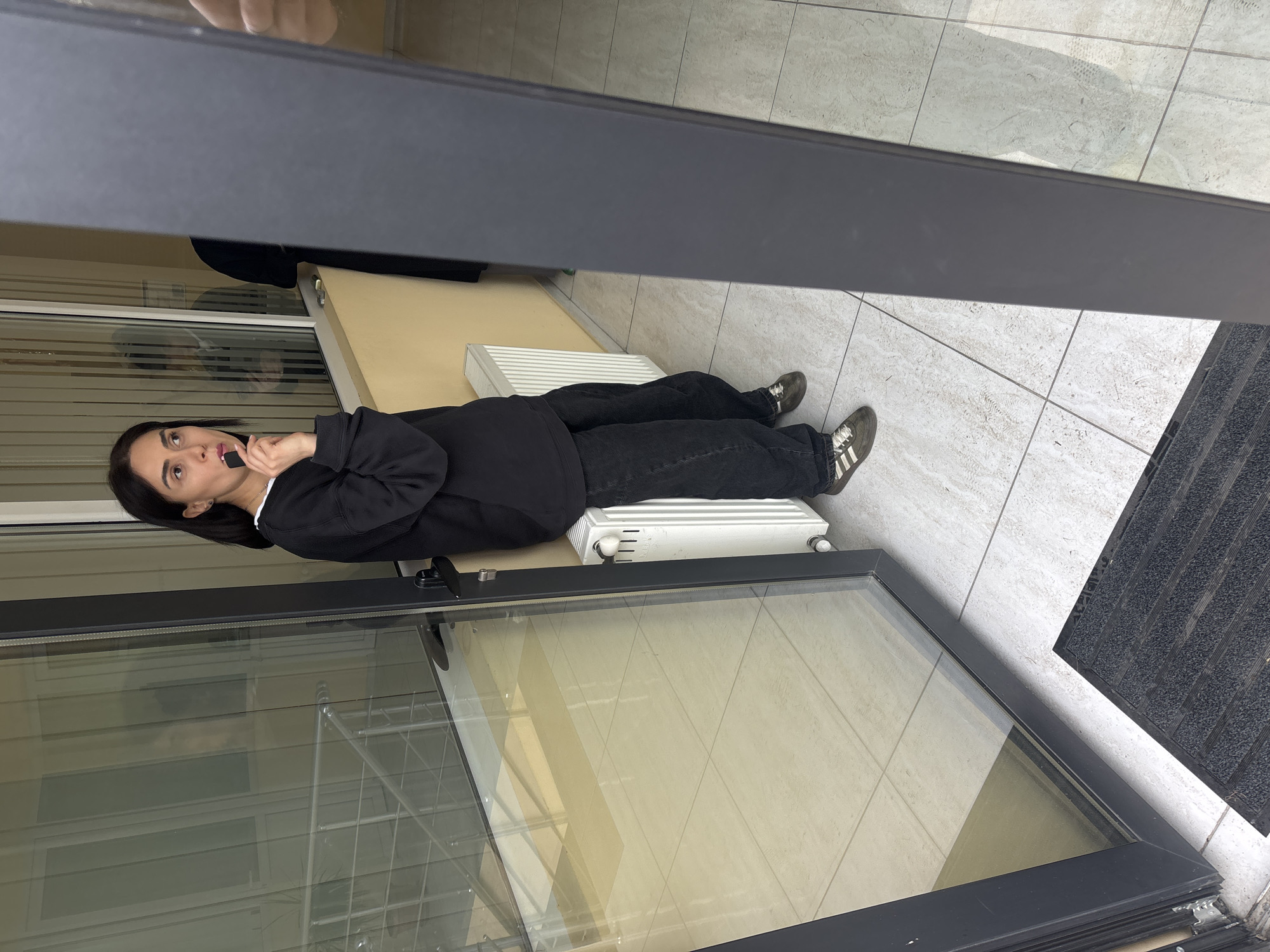 The image depicts an adult female positioned within an indoor common area of a building in Tbilisi, Georgia. The woman has dark, shoulder-length hair and is dressed in a black long-sleeved sweatshirt with a white collar visible underneath, dark baggy trousers, and olive green Adidas sneakers with white stripes. She is standing with her back leaning against a white panel radiator, which is mounted on a light yellow wall beneath a window. Her right hand holds a small, dark object, resembling a vape pen or lip product, to her lips. Her head is tilted slightly, and her gaze is directed upwards and to her left.
To the woman's left, a large glass partition reflects an interior staircase with metal railings and a faint, dark silhouette of another individual. The floor throughout the visible area is composed of light-colored tiles. A dark grey door frame partially obstructs the right side of the frame, and a black ridged doormat is visible on the floor in the lower left foreground, indicating a threshold.