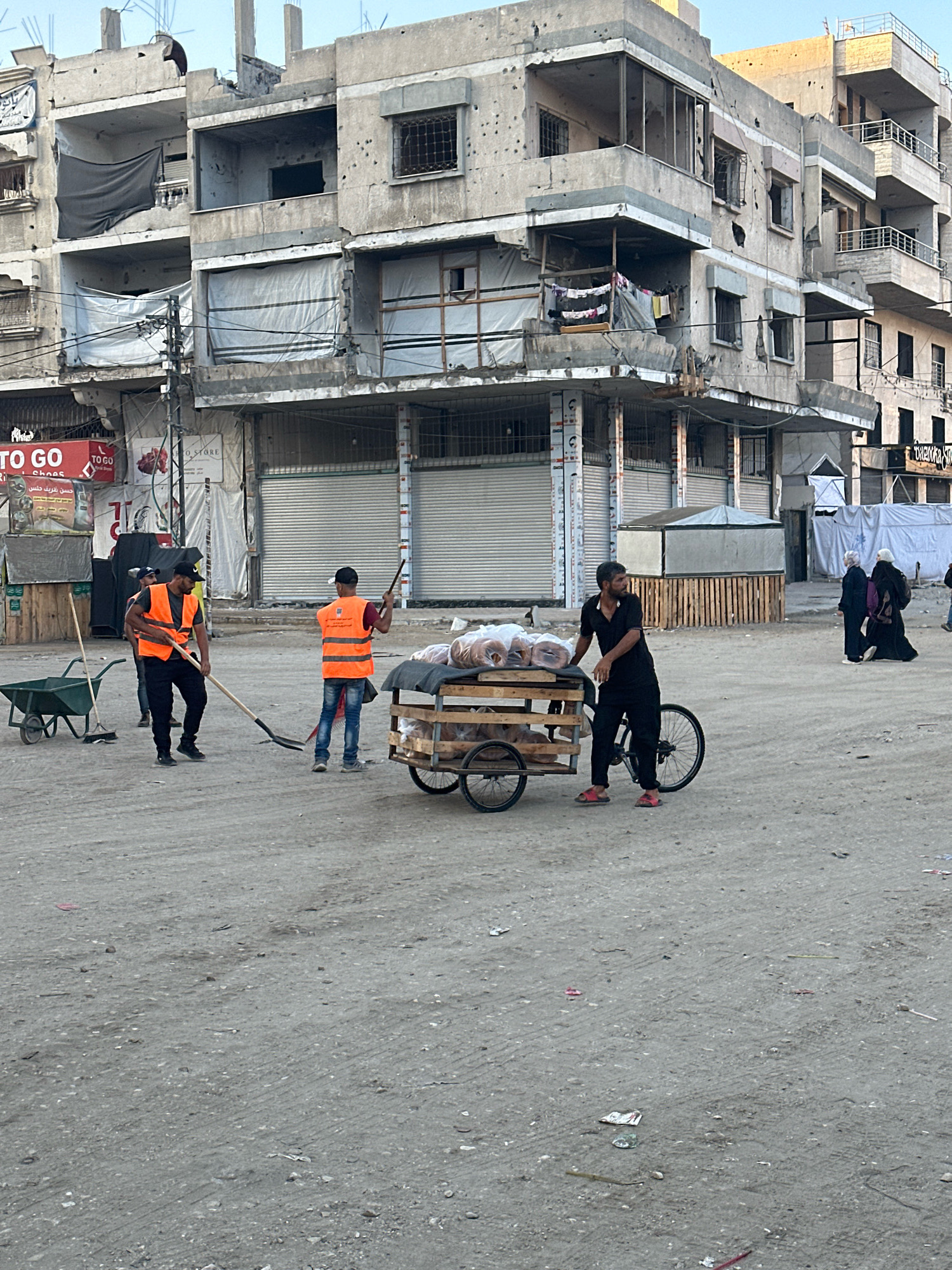In the city of Gaza, Palestinian Territories, a street scene unfolds amidst the remnants of damaged buildings. The facades of multi-story structures bear the marks of conflict, with visible bullet holes and debris. Several windows are covered with tarps or shutters, and some balconies display laundry.
In the foreground, several individuals are engaged in activity. Two men in high-visibility orange vests and hard hats are working, one with a shovel and the other with a broom, suggesting cleanup efforts. Nearby, a man in a black shirt and pants pushes a wooden cart filled with what appear to be rolls of bread, attached to a bicycle frame. He is wearing red sandals. Further in the background, two women in hijabs are walking away from the camera, one carrying a purple bag.
The ground is uneven and dusty, characteristic of a street in a state of disrepair. A green wheelbarrow and other discarded materials are visible. The overall atmosphere suggests a community grappling with recovery and daily life in a challenging environment.