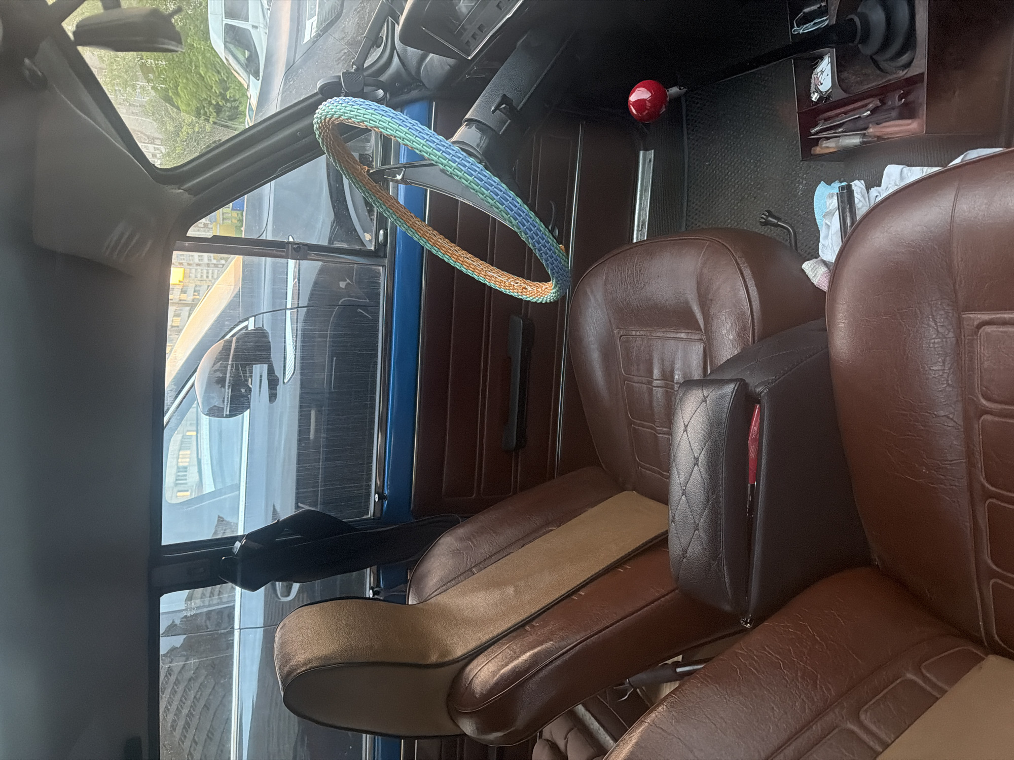 The image provides an interior view of a car, likely a vintage model given the design of the seats and dashboard. The seats are upholstered in brown leather with a diamond-stitched pattern and a contrasting tan fabric insert on the driver's seat headrest. A black leather armrest with a diamond-stitched pattern sits between the front seats. The steering wheel is covered with a crocheted multicolor cover in shades of green, blue, and orange. A bright red shift knob is visible on the center console. The dashboard appears dark and somewhat worn, with a silver-trimmed instrument cluster. The floor is covered with a black textured mat. Through the windows, other vehicles and buildings can be seen, suggesting the car is parked on a street or in a parking lot in an urban environment. The lighting indicates it might be late afternoon or early evening. The scene is devoid of any people.
