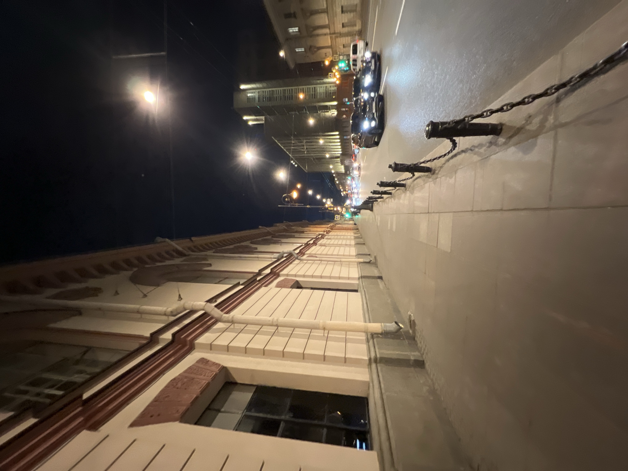 This is a night scene on a street in Saint Petersburg, Russia. The view is from a low angle, possibly street level.
The right side of the image depicts a sidewalk with a low concrete wall adorned with cannon-shaped bollards connected by chains. A street runs parallel to the sidewalk, with cars visible.
The left side of the image shows a building facade with windows and decorative elements. Street lights illuminate the scene, with several visible in the distance. The sky is dark.