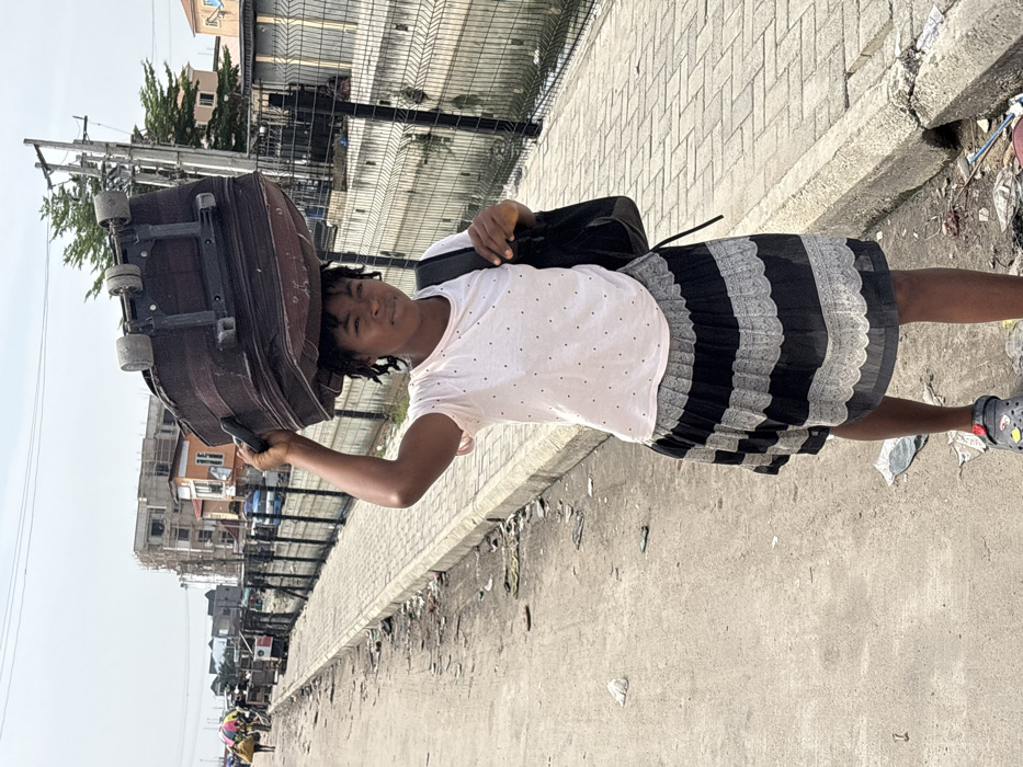 A young female, appearing to be in her late teens or early twenties, walks on a dirt pathway in the city of Igboefon, Nigeria, under a bright, overcast sky. She is centrally positioned, facing the viewer directly. She has dark skin and braided hair, and is dressed in a light pink t-shirt with dark polka dots, a black and grey pleated skirt with a lace-like pattern, and a single grey Croc-style shoe on her visible left foot. She carries a dark brown, worn wheeled suitcase balanced on her head, stabilized by her raised right hand. A black shoulder bag hangs from her left shoulder. To her immediate right, a raised grey brick-paved sidewalk parallels a metal mesh fence, behind which runs a narrow, open canal. The dirt pathway she traverses is littered with various debris. In the background, additional individuals are visible further down the path, and an urban landscape unfolds with multiple buildings, including one under construction, and utility poles with overhead lines.