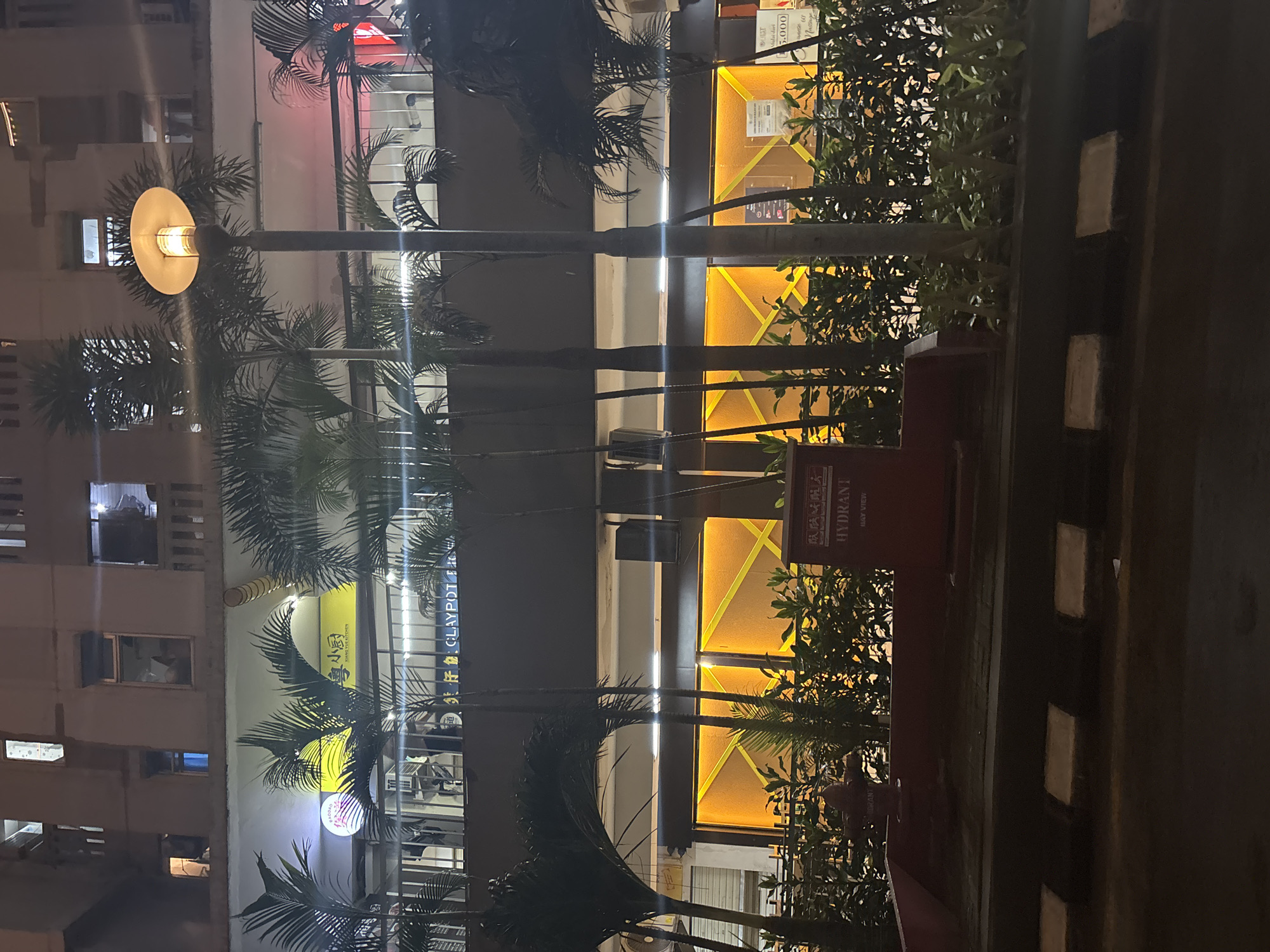 The image depicts a nighttime street view of a commercial building with multiple levels. The facade features several windows and balconies on the upper floors. Below, a row of palm trees partially obscures a storefront with illuminated signage in yellow and white, indicating "XIANG YUE KITCHEN" and "CLAYPOT". Further down, a red hydrant is visible in the foreground, surrounded by greenery. The street in front has striped markings. The overall scene suggests an urban environment in North Jakarta, Indonesia, likely a commercial area at night.