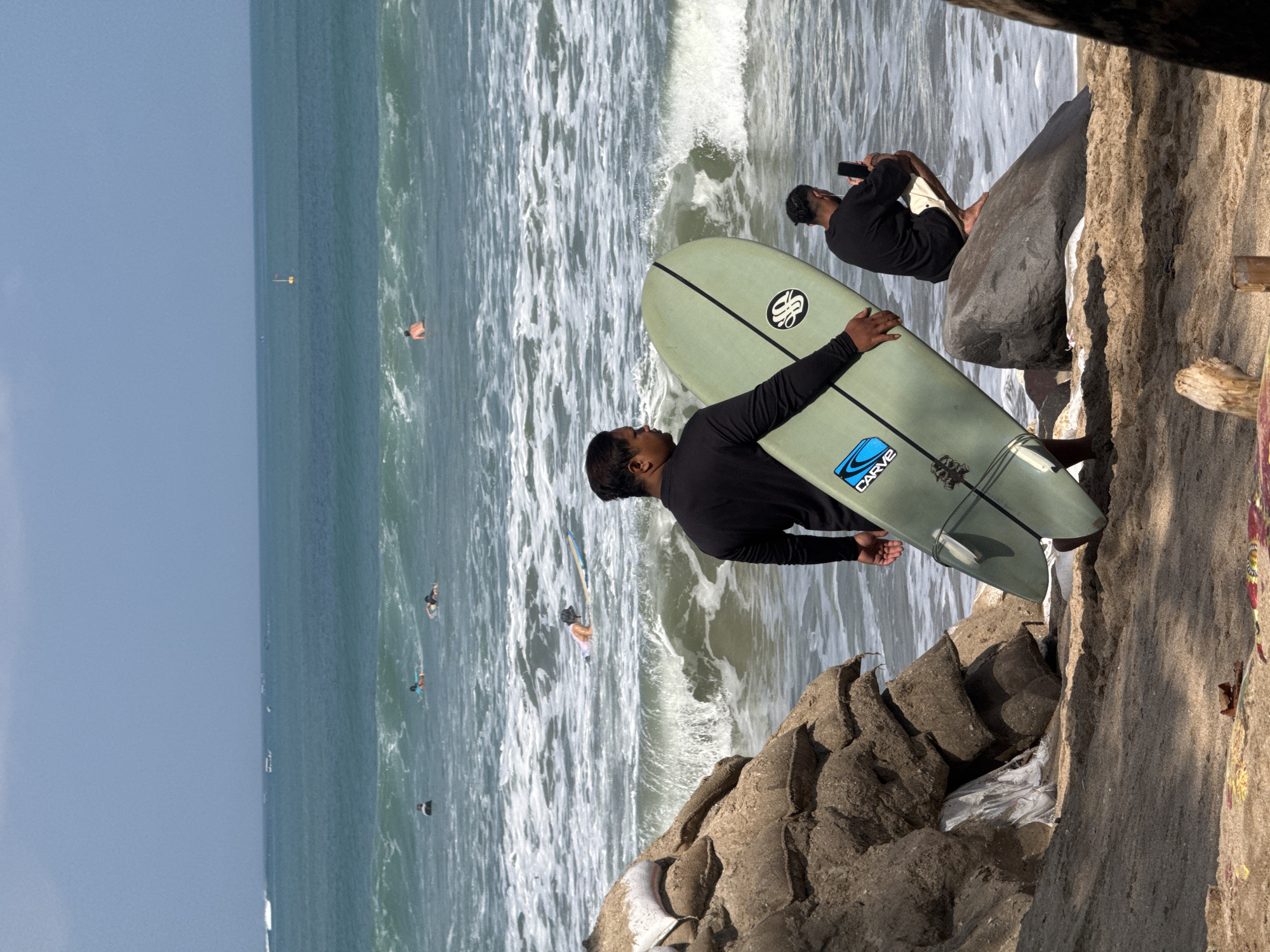 The image depicts a beach scene in Kuta, Indonesia, with the ocean in the background and a sandy shore in the foreground. A man in a black long-sleeved shirt is holding a light green surfboard, standing on the sand. To his right, another man wearing a black t-shirt and light-colored shorts is crouched on a large rock by the water, looking at his phone. Several surfers are visible in the turquoise water, some on surfboards and others swimming. The waves are moderate, with white foam breaking near the shore. On the left side of the image, a pile of sandbags is stacked up, partially covering some white plastic sheeting. The sky is overcast and hazy.