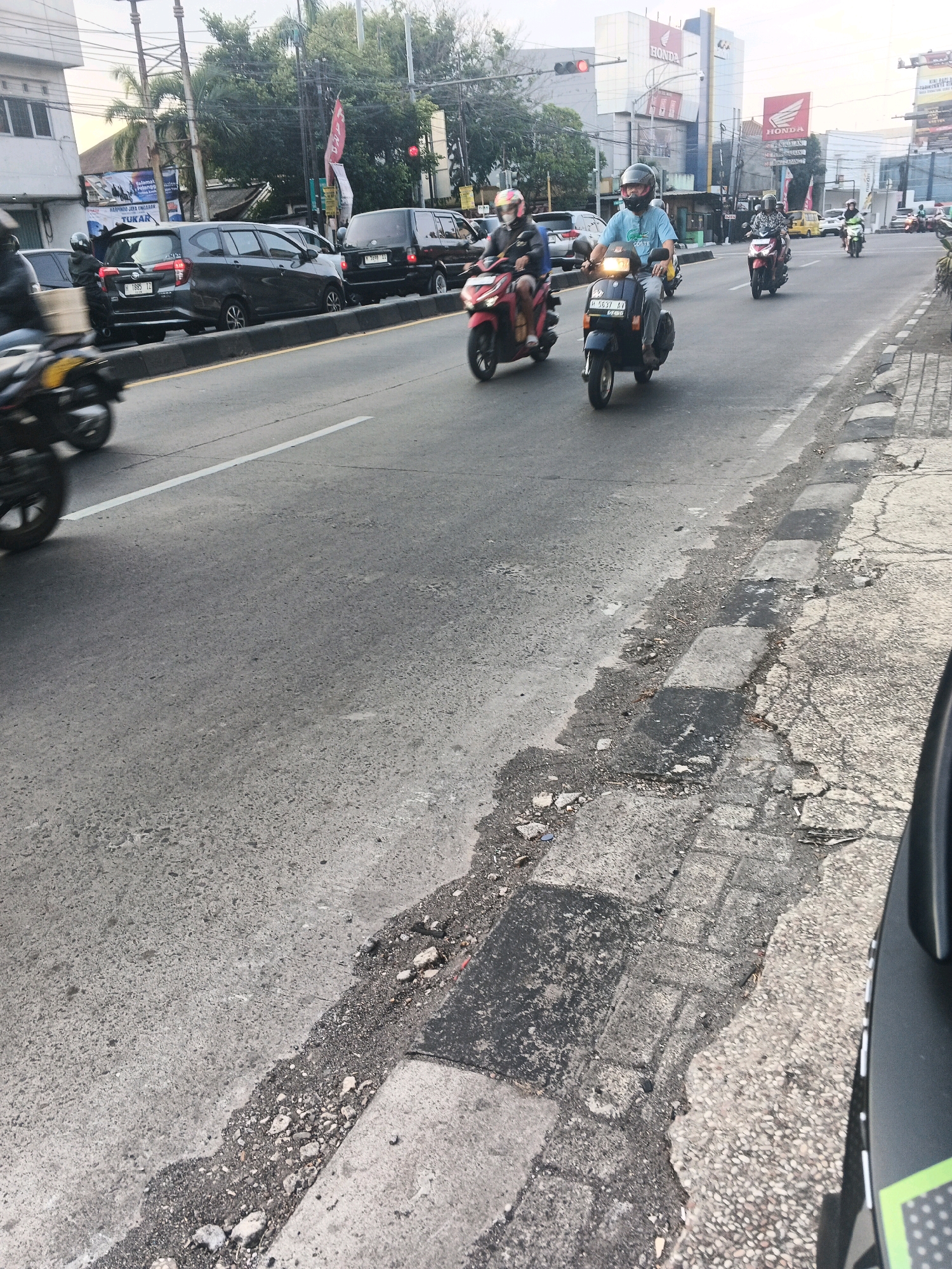 The image shows a busy street scene in Ungaran, Indonesia. Several motorbikes and cars are on the road, with some motorbikes being ridden by people wearing helmets. A large grey car is parked on the side of the road, and a red and white banner with text is visible in the background. The road appears to be well-trafficked, and the buildings in the background suggest an urban environment. There is also a pedestrian walkway with visible cracks and debris. The overall impression is one of a typical day on a main road in an Indonesian city.