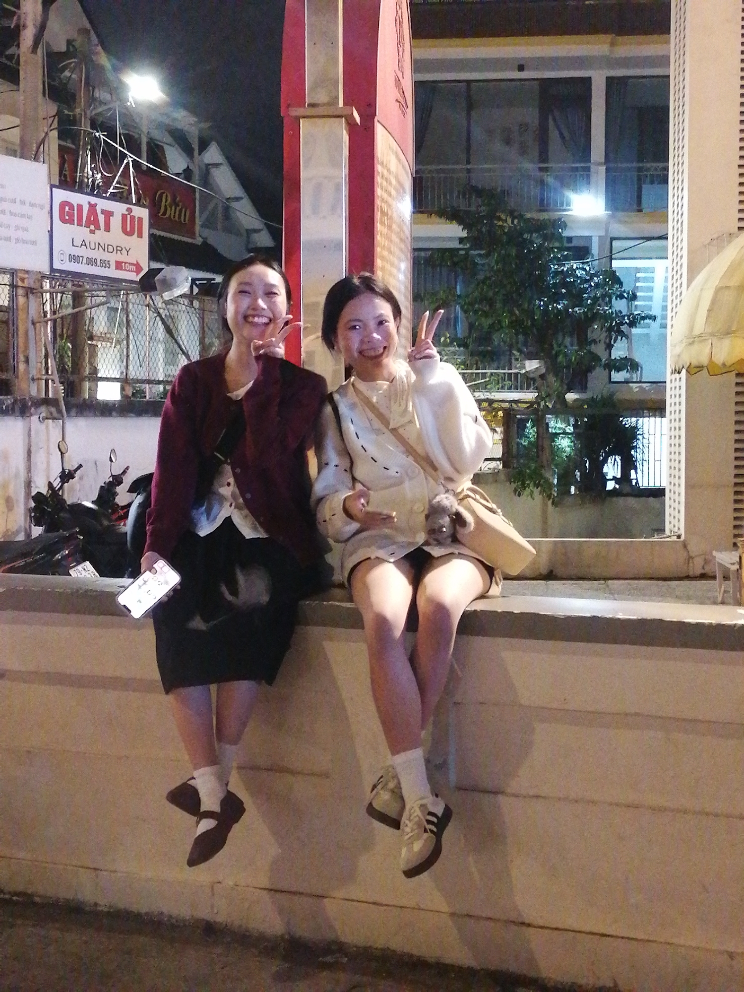 Two young women are sitting on a concrete ledge outdoors at night. The woman on the left has dark hair, is wearing a maroon cardigan over a white top and a black skirt. She is holding a smartphone and has one hand raised with two fingers extended in a peace sign. She is smiling broadly. The woman on the right has dark hair and is wearing a cream-colored sweater with voluminous sleeves and a light-colored skirt. She is also making a peace sign with her fingers and smiling. She has a light-colored crossbody bag with a fluffy charm attached. Both women are wearing white socks and dark shoes.
Behind them, a building with multiple floors and windows is visible, along with trees and a street light. A sign with Vietnamese text, "GIẶT ỦI LAUNDRY" and a phone number, "0907.069.655" is visible on the left side of the frame, along with a motorcycle parked behind it. The scene appears to be in an urban or semi-urban setting. The lighting suggests it is nighttime. The overall mood is cheerful and relaxed.