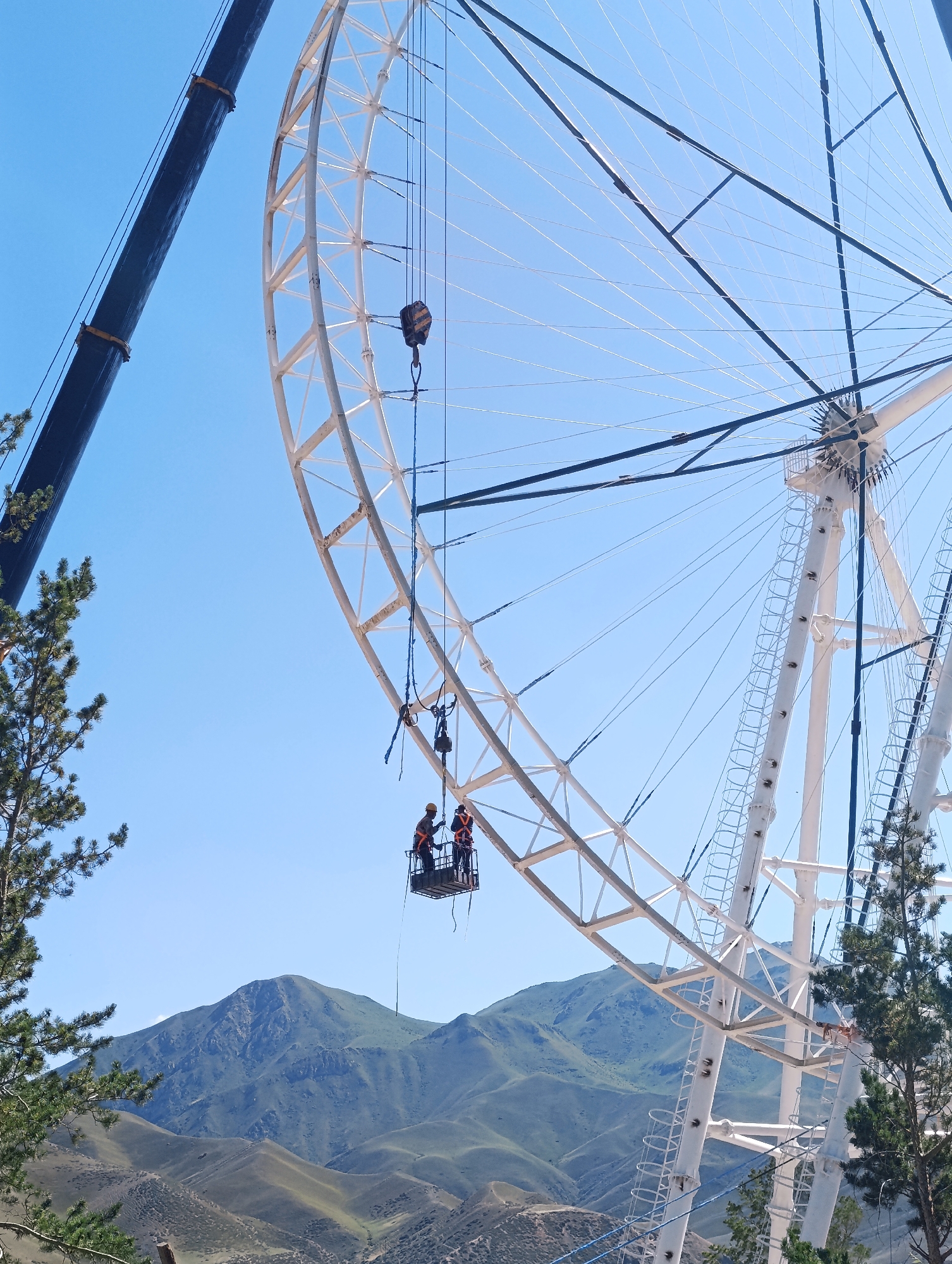 In Naryn City, Kyrgyzstan, a large Ferris wheel is under construction or maintenance, with two workers in a suspended gondola near the top of its white metal frame. They are wearing safety harnesses and helmets. A large crane boom is visible in the upper left corner of the image, extending towards the Ferris wheel. The background features a clear blue sky and rolling, green, mountainous terrain. A pine tree is in the foreground on the left, and another on the right. The scene appears to be outdoors during daylight hours.