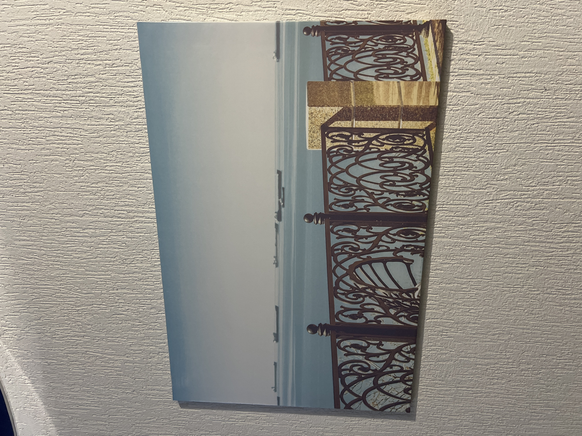 The image depicts a panoramic canvas print of a seascape, likely a waterfront scene in Yeysk, Russia, prominently displayed on a textured white wall. The artwork itself features a calm, light blue ocean under a hazy sky, with a line of several large cargo ships visible on the horizon. In the foreground, a decorative, dark metal fence with ornate scrollwork runs across the width of the canvas. To the right of the fence, a rectangular stone pillar stands, casting a shadow. The overall impression is one of a serene, possibly historical, coastal view. The wall on which the print is hung has a distinct, vertical, ridged texture.