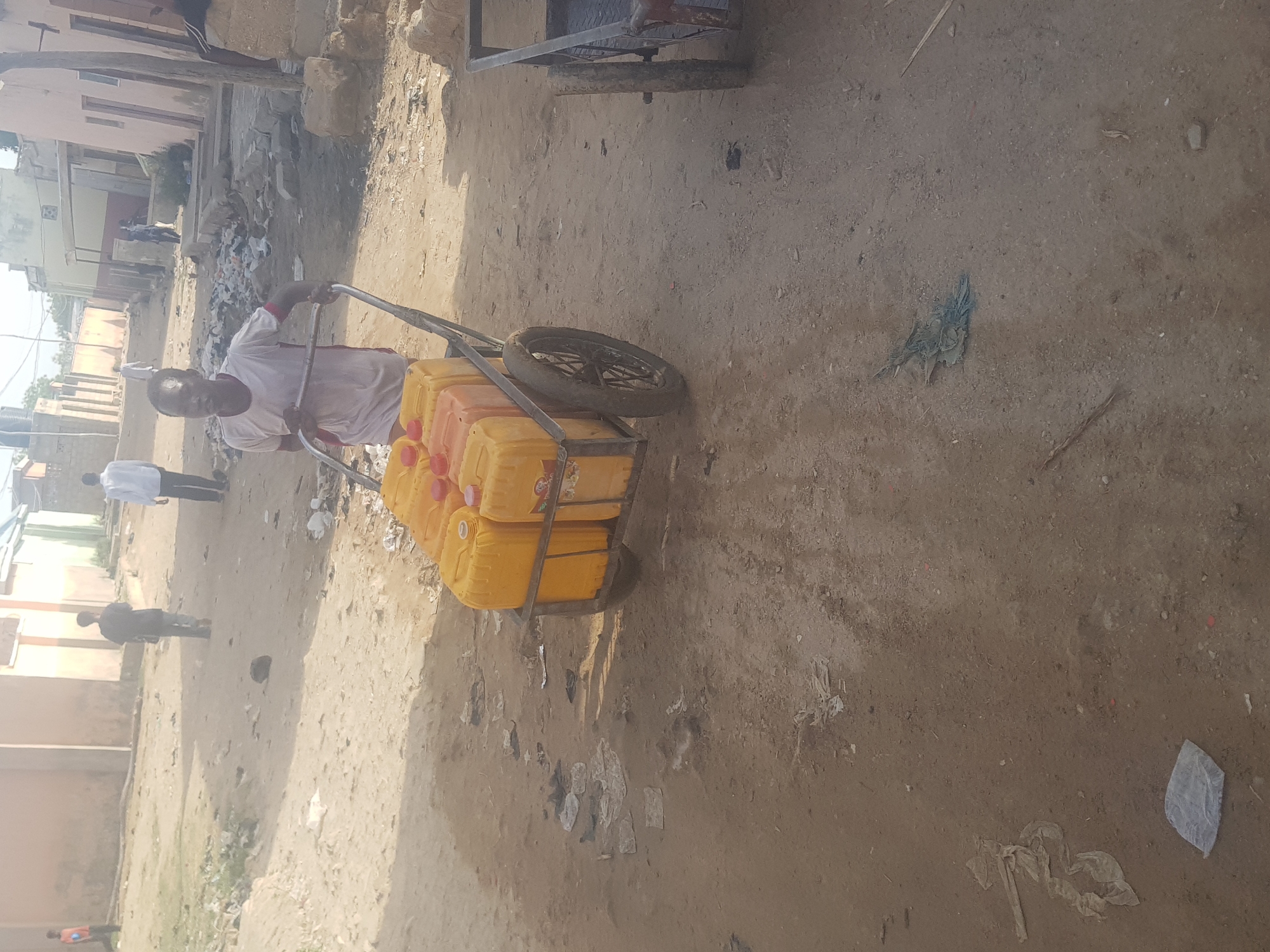 The image shows a man pulling a cart loaded with yellow jerrycans in Dabawa, Nigeria. The man is wearing a white shirt and appears to be older. The cart has one wheel and a metal frame holding the jerrycans, which are likely filled with water. The ground is dirt and dusty, with scattered trash and debris. Other people are visible in the background, walking along the dirt road. Buildings and structures, possibly homes or shops, line the street. The scene conveys a sense of a basic and possibly impoverished environment, where transporting water is a manual and visible task.