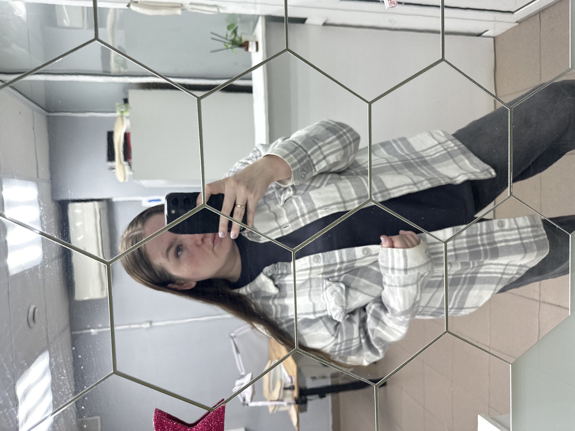 The scene, located in Chaikovsky, Russia, depicts an adult female capturing a mirror selfie within an indoor commercial or office environment. The woman, with long brown hair, wears an oversized light gray and white plaid button-up shirt over a solid dark gray or black crew-neck top, paired with dark gray trousers. Her right hand, adorned with a gold-colored ring on the ring finger and light-colored nail polish, holds a dark smartphone (likely an iPhone due to the visible camera array) towards the mirror. Her left hand rests on the lower front of her plaid shirt.
The mirror itself is constructed from multiple hexagonal tiles, separated by thin, light-colored metallic lines. Reflected in the mirror, the background reveals a room with light gray walls and a beige-tiled floor. Overhead, fluorescent lighting fixtures are visible on the ceiling, along with a circular white smoke detector and two white wall-mounted air conditioning units. To the left, a partial pink glittery star decoration is visible, alongside a wooden table with a black base, a flexible-arm desk lamp, and a small white container. To the right, a white shelf or counter displays a small brown bottle with diffuser sticks and a green potted plant. A white wall-mounted telephone handset and part of a white door frame are also visible on the far right.