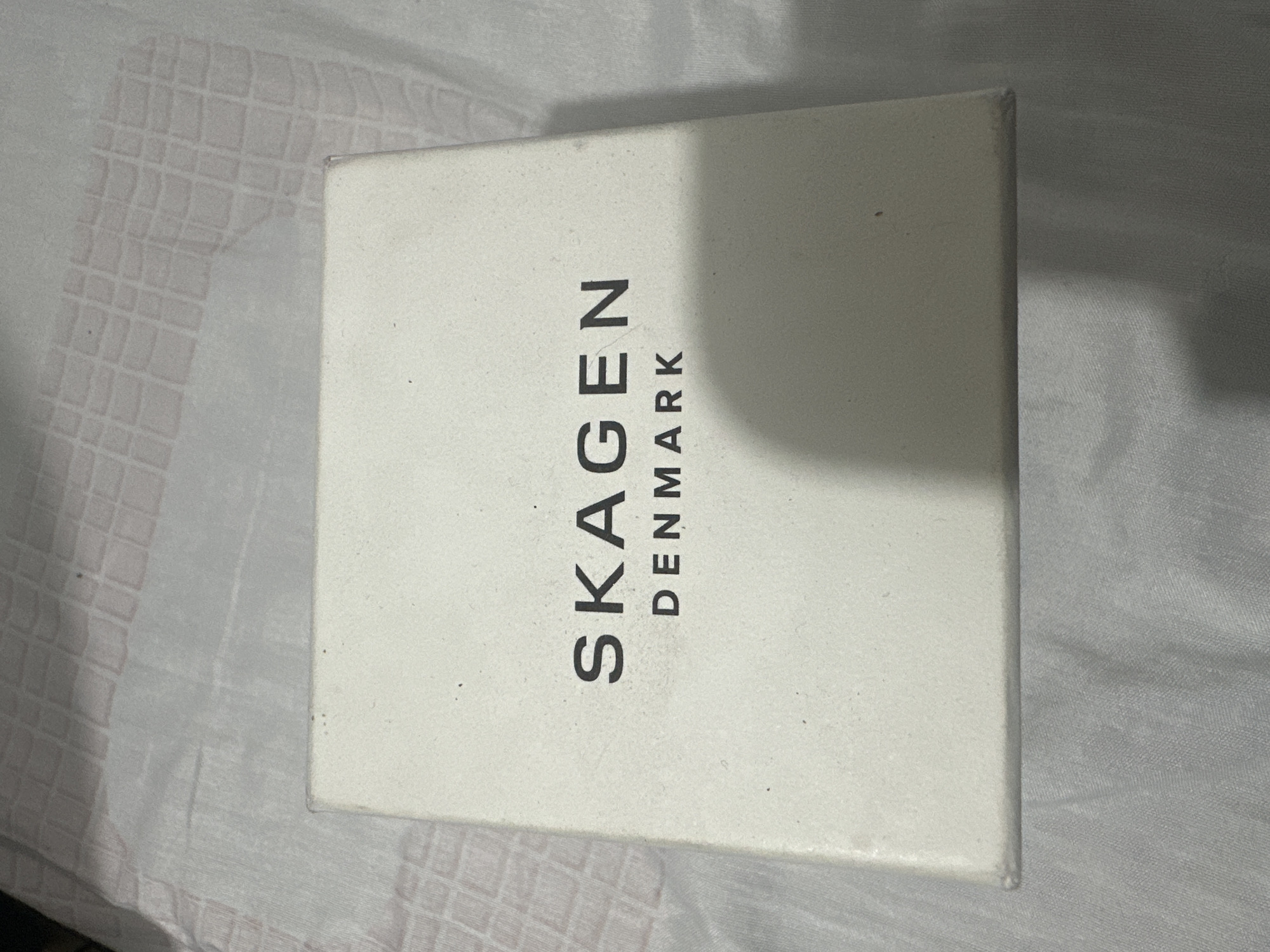 The image shows a white box with "SKAGEN DENMARK" printed on it. The box is placed on a textured, light-colored surface, possibly fabric. The lighting is dim, and there are shadows cast on the surface. The image quality is slightly grainy, and there are a few small dark specks on the box. No specific location within Davao City, Philippines, can be determined from the visual information provided.