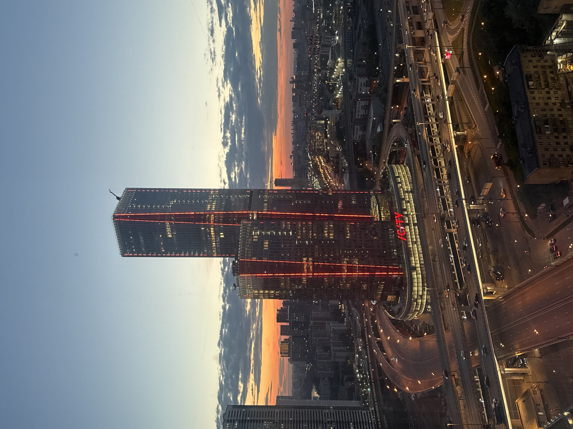 A high-angle, vertical shot captures a bustling urban cityscape at dusk or sunset, with prominent modern architecture and an intricate road network. The scene is set in Khimki, Russia.
The primary subjects are two tall, contemporary skyscrapers, distinguished by their dark facades and striking red illuminated lines that run vertically along their exteriors. The shorter of these two towers features the red text "iCITY" prominently displayed on its side. In the background, stretching towards the horizon, is a sprawling urban area with numerous other buildings of varying heights.
Below the skyscrapers, a complex system of multi-lane highways and elevated roadways is visible, bustling with traffic. Numerous cars, identifiable by their headlights and taillights, navigate the well-lit roads and overpasses. Streetlights brightly illuminate the asphalt, creating glowing trails and patterns.
The time of day is clearly twilight, as indicated by the vivid sunset or sunrise colors on the horizon—a gradient of warm oranges and yellows blending into a cooler blue sky, interspersed with clouds. The artificial lights of the city are fully active, contrasting with the fading natural light. The overall atmosphere conveys a sense of a modern, active metropolis. No specific human figures or distinct activities beyond traffic are visible.