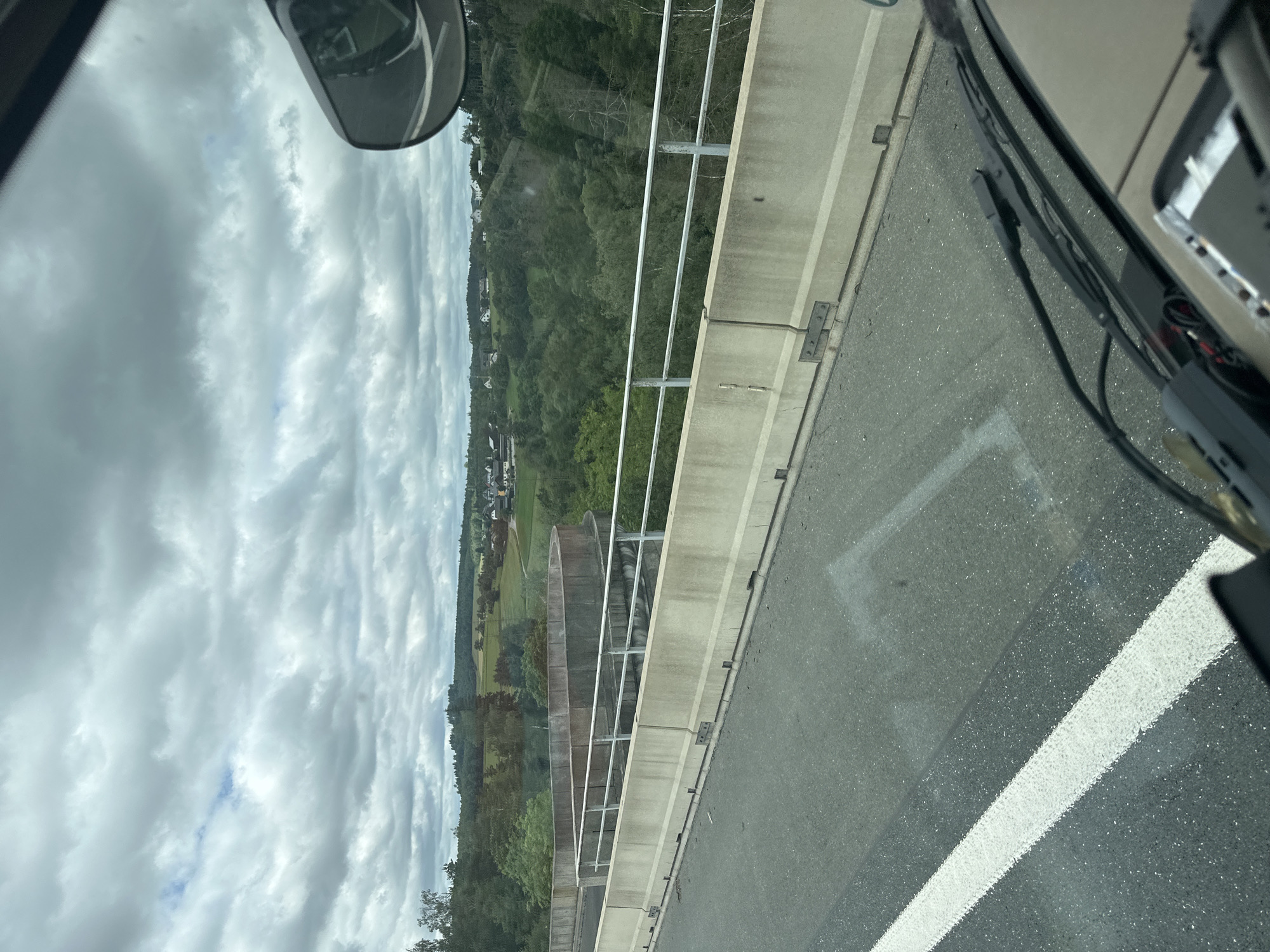 The image is taken from the perspective of a vehicle driving on a highway overpass in Köditz, Germany. The highway has a grey asphalt surface with a white lane marking on the left side. The overpass is bordered by concrete barriers and a metal railing. Beyond the overpass, a lush green valley with scattered houses and buildings is visible. The sky is overcast with dramatic grey clouds. The overall scene depicts a rural landscape in Germany seen from a moving vehicle.