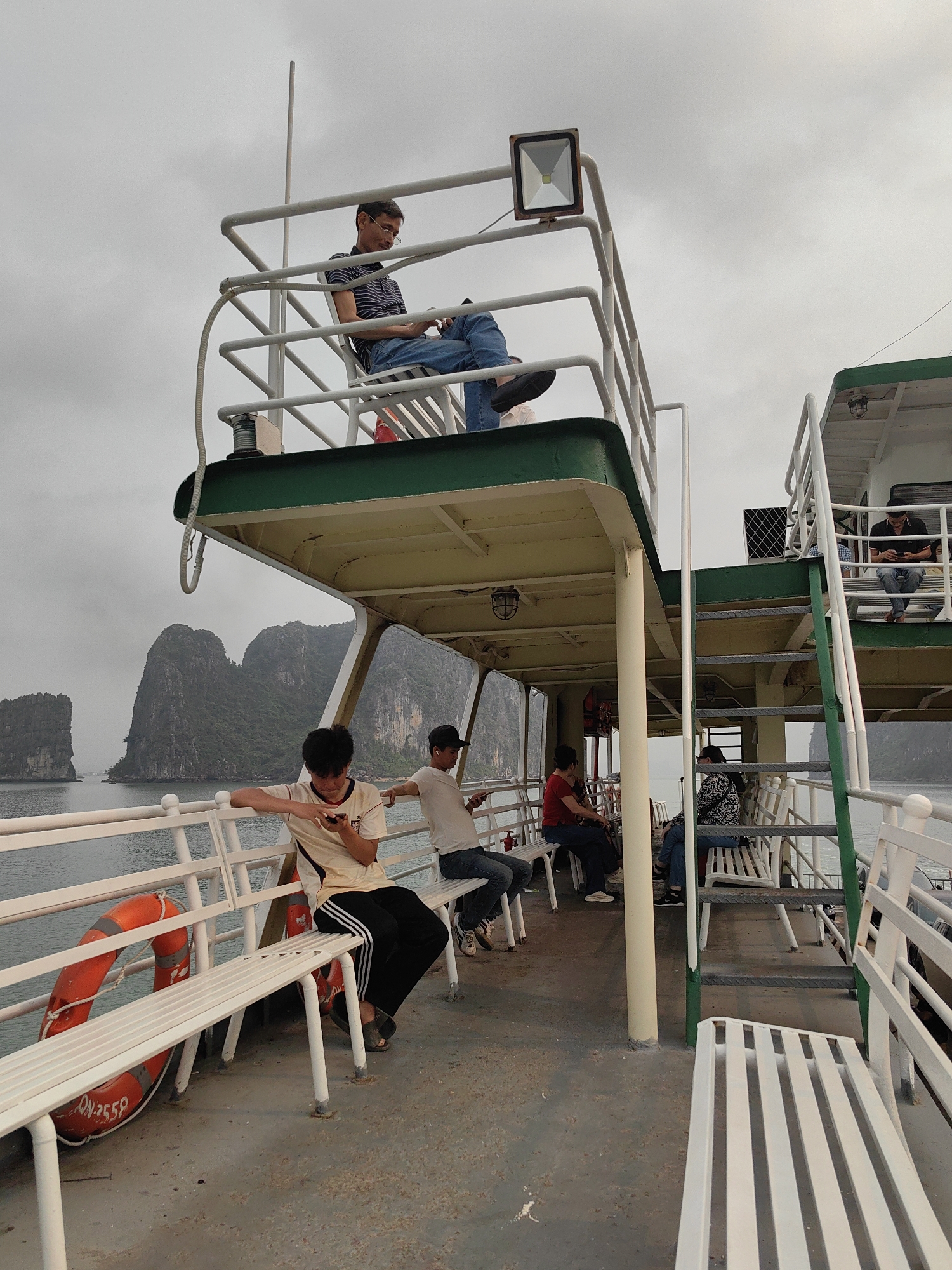 On a boat in Ha Long Bay, Vietnam, under an overcast sky, several people are scattered on the deck. A man sits on a raised platform looking at his phone. Below, a young man in a white shirt with black stripes and black pants leans against the railing looking at his phone. Near him, another man in a white t-shirt sits on a bench, also looking at his phone. Other passengers are seated on benches. A life preserver is attached to the railing. Rock formations are visible in the water in the background.